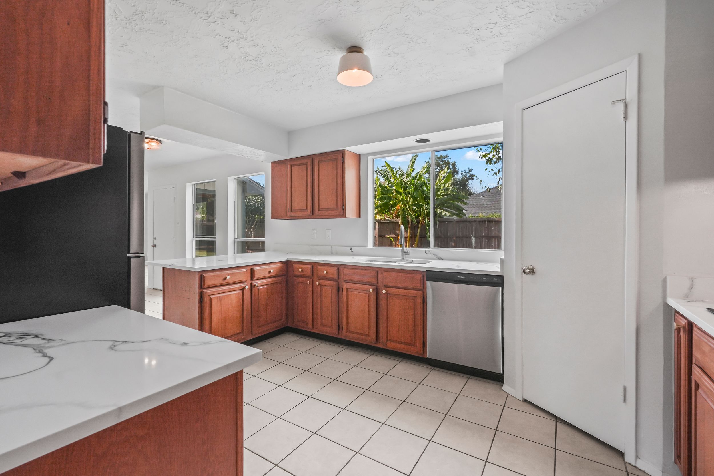 Spacious kitchen with cherry wood cabinets, marble countertops, tile flooring, stainless steel dishwasher, and bright windows.