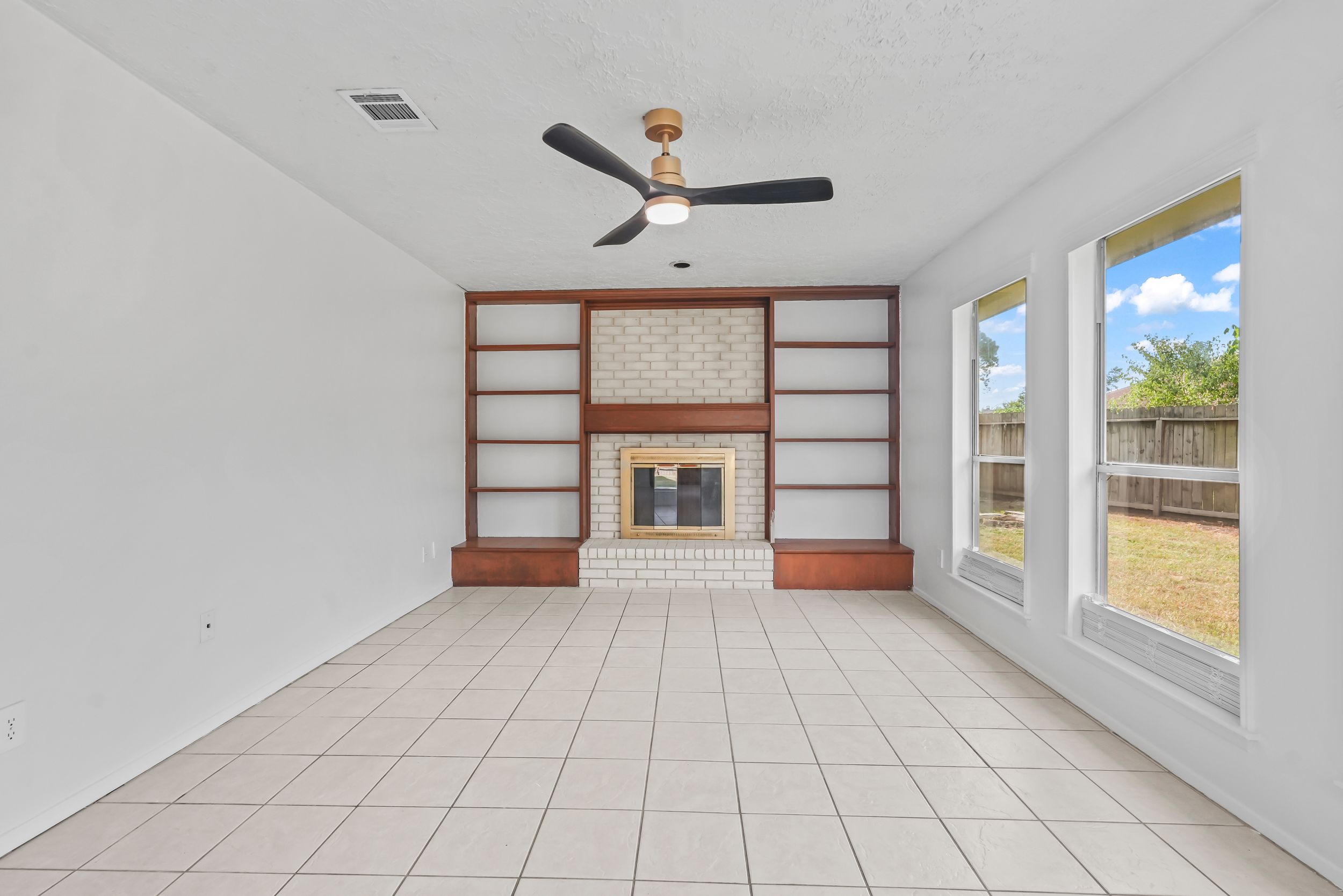 Spacious living room with tile floors, brick fireplace, built-in shelves, ceiling fan, and large windows with backyard view.