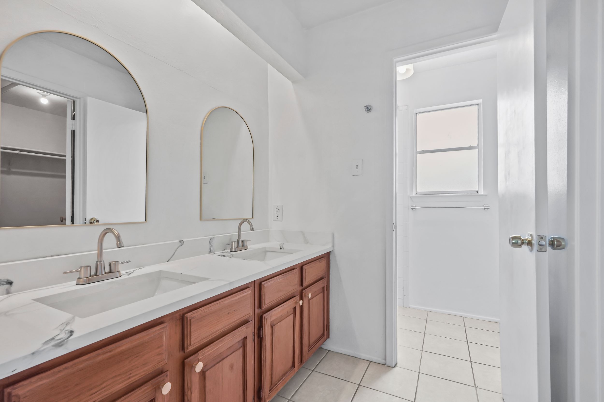 Bright bathroom with double sink vanity, wood cabinets, white countertops, arched mirrors, and tile flooring.