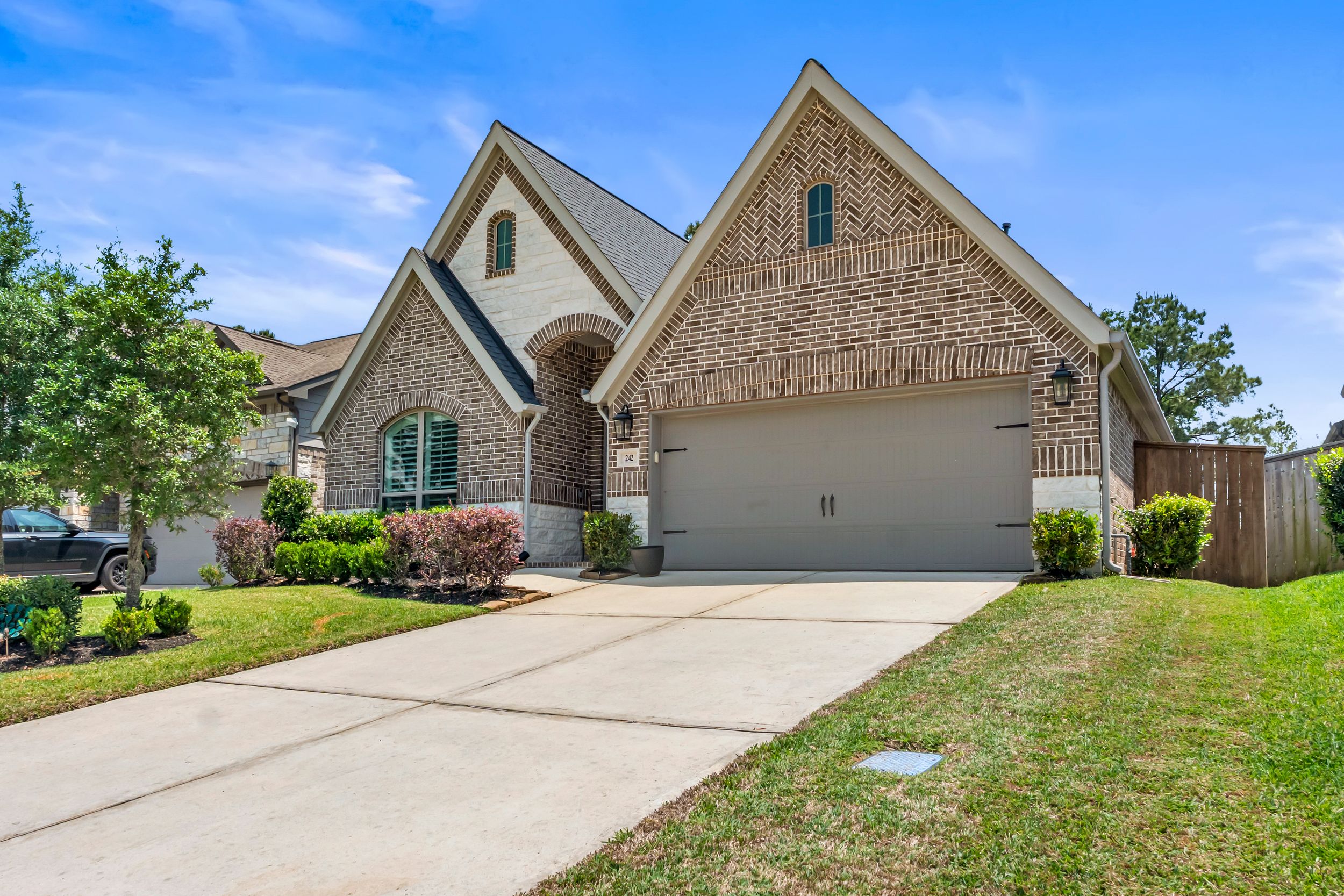 Brick two-story home with arched entry, two-car garage, manicured lawn, and lush trees on a sunny day.