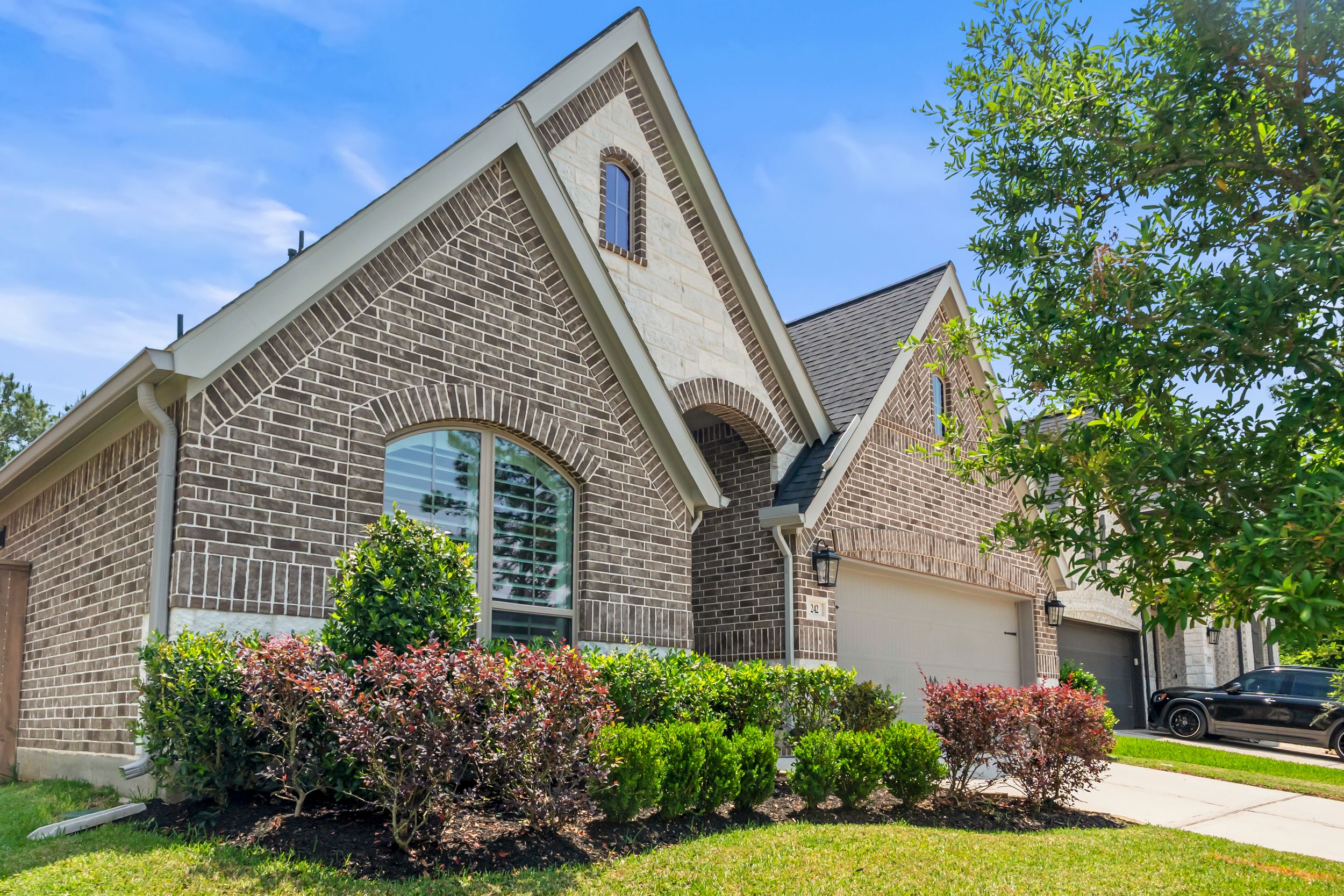 Brick two-story home with arched windows, two-car garage, and lush green landscaping under a bright blue sky.