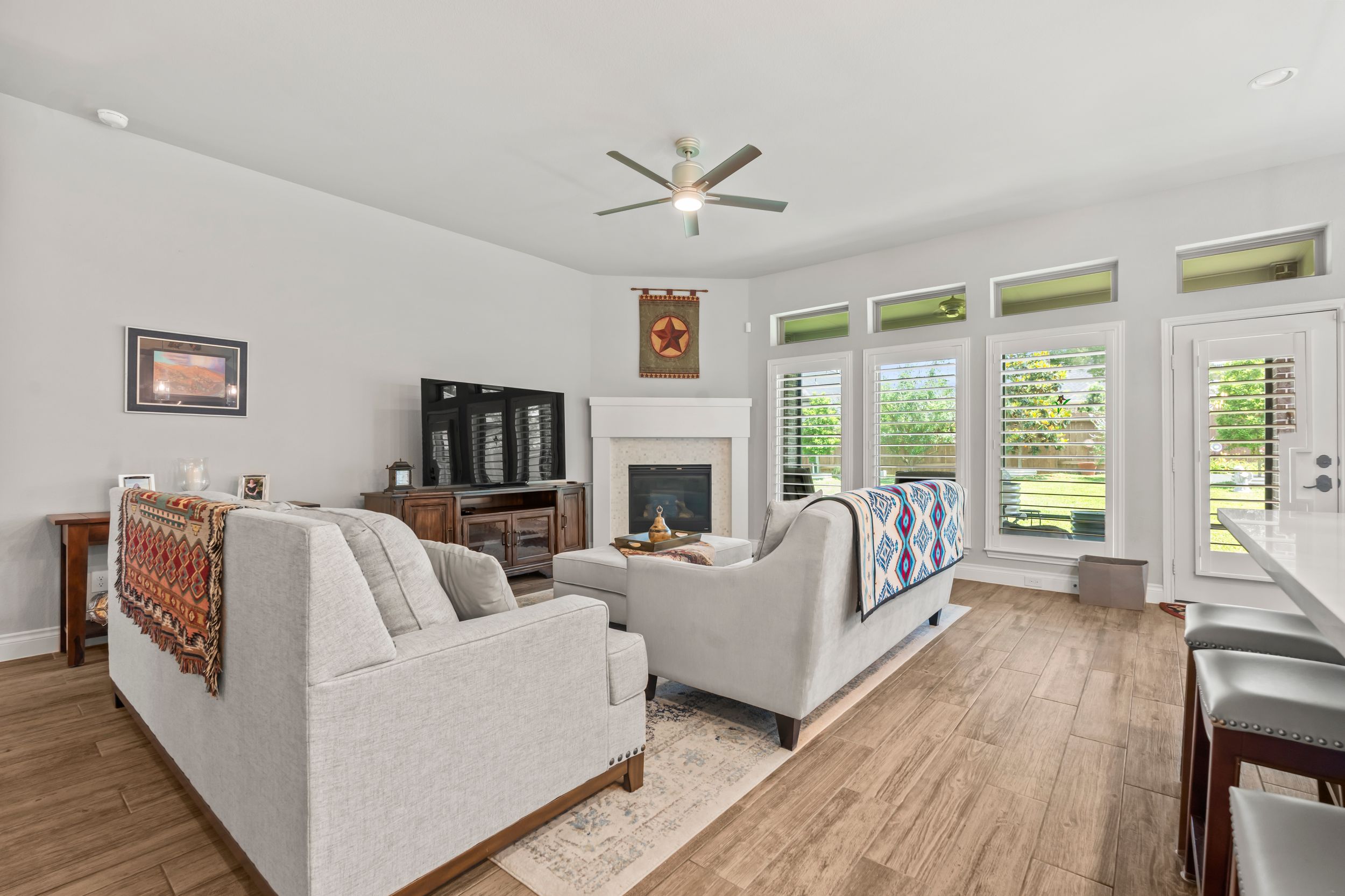 Bright living room with gray sofas, hardwood floors, fireplace, and ceiling fan with large windows.