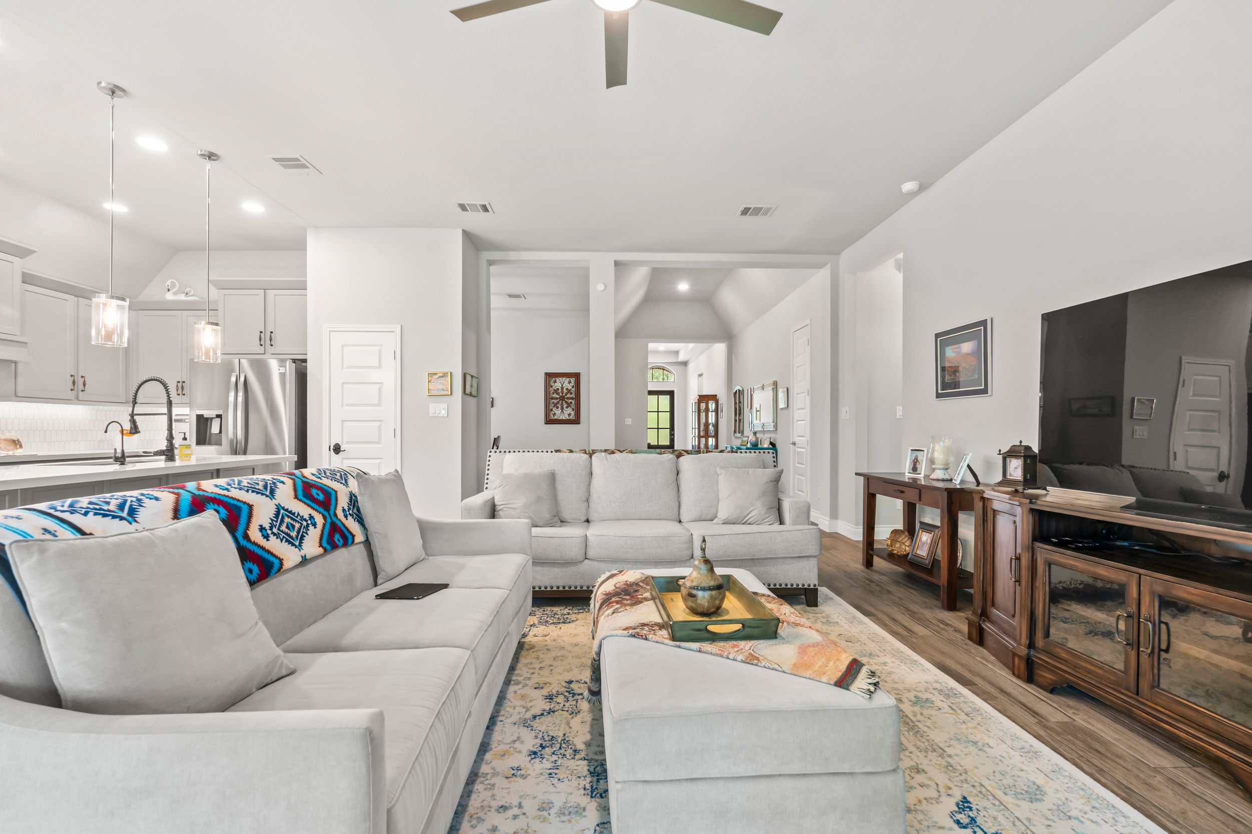 Bright open-concept living room with gray sectional sofa, ceiling fan, hardwood floors, and modern kitchen in background.