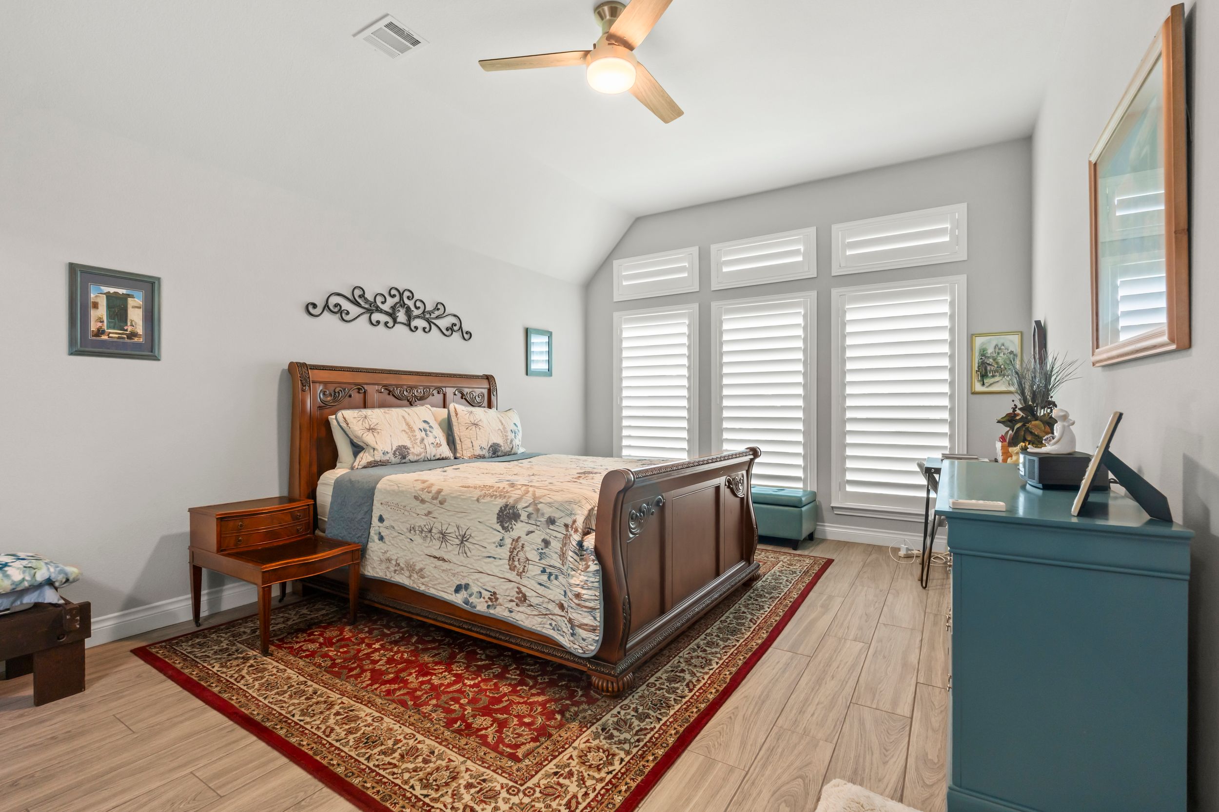 Bright bedroom with vaulted ceiling, ceiling fan, wood sleigh bed, red area rug, plantation shutters, and light hardwood floors.