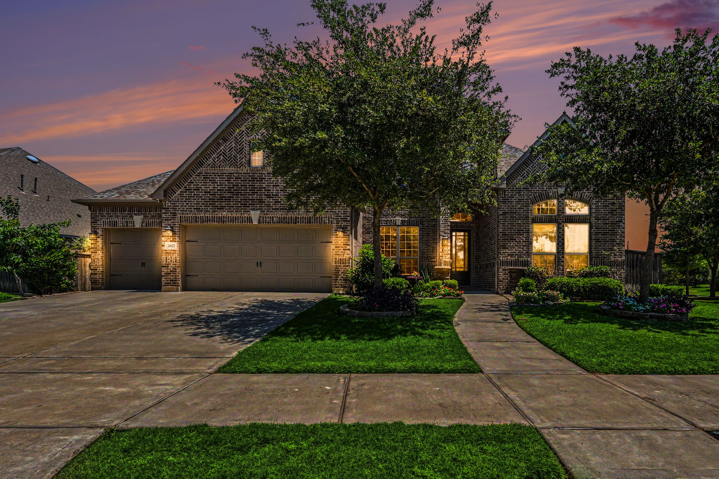 Brick home with two-car garage illuminated at dusk with lush green lawn and mature trees under a colorful sunset sky.