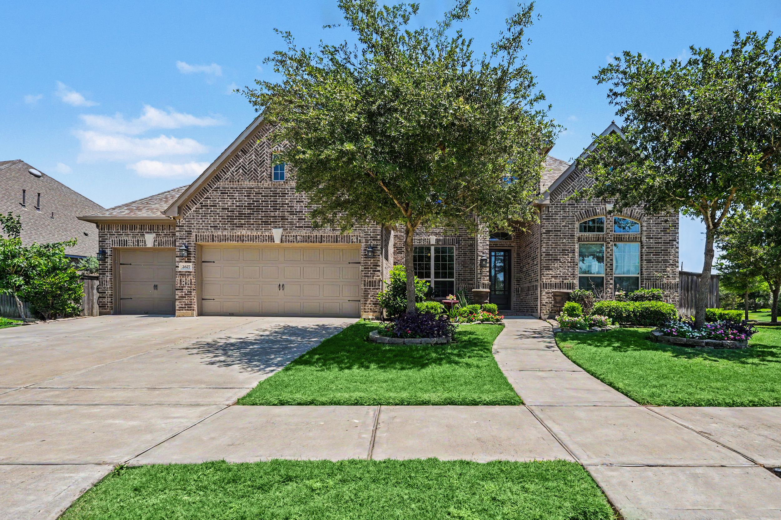 Brick single-story home with two-car garage, lush green lawn, mature trees, and manicured landscaping on a sunny day.