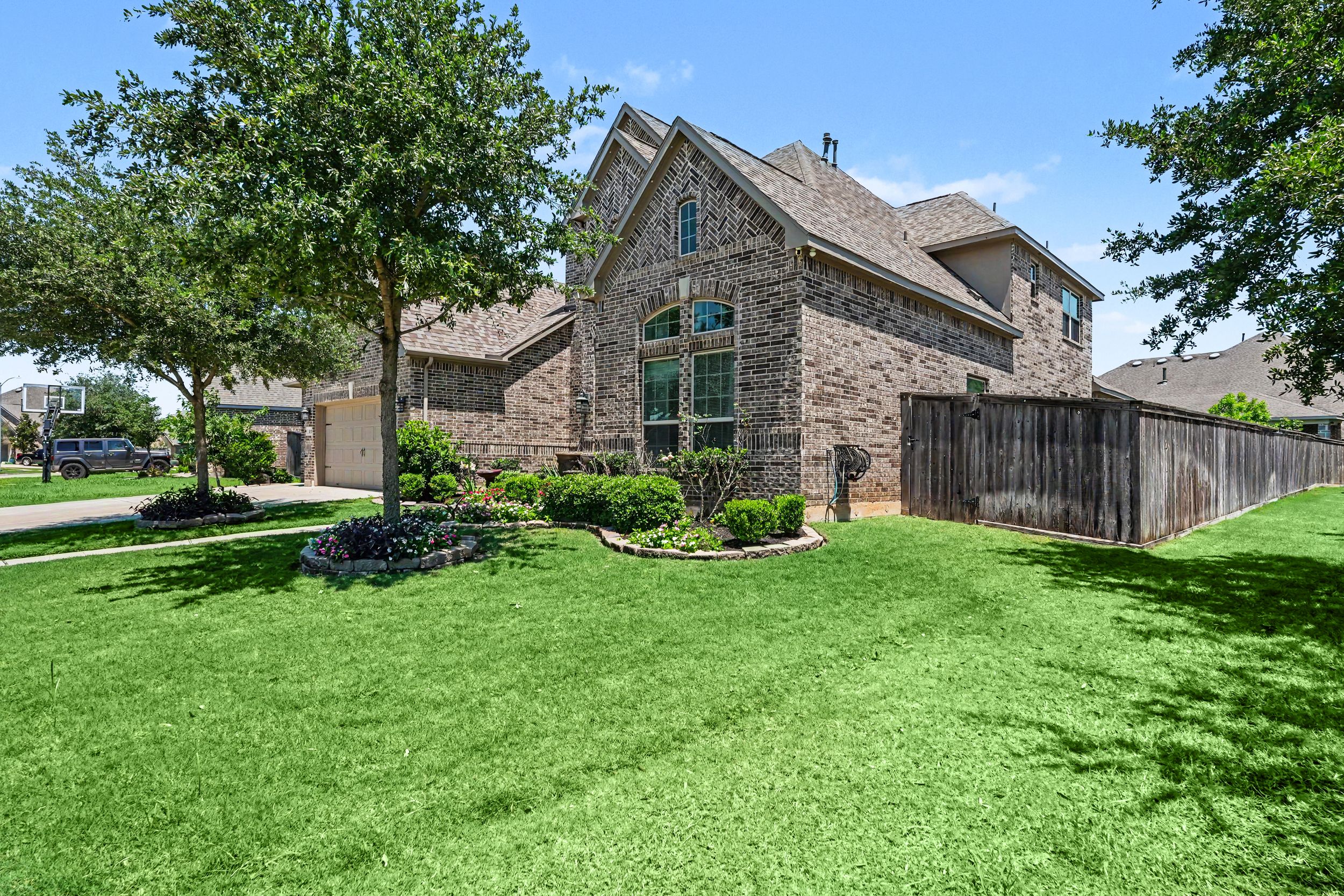 Two-story brick home with large green lawn, mature shade trees, and wooden privacy fence on a sunny day.