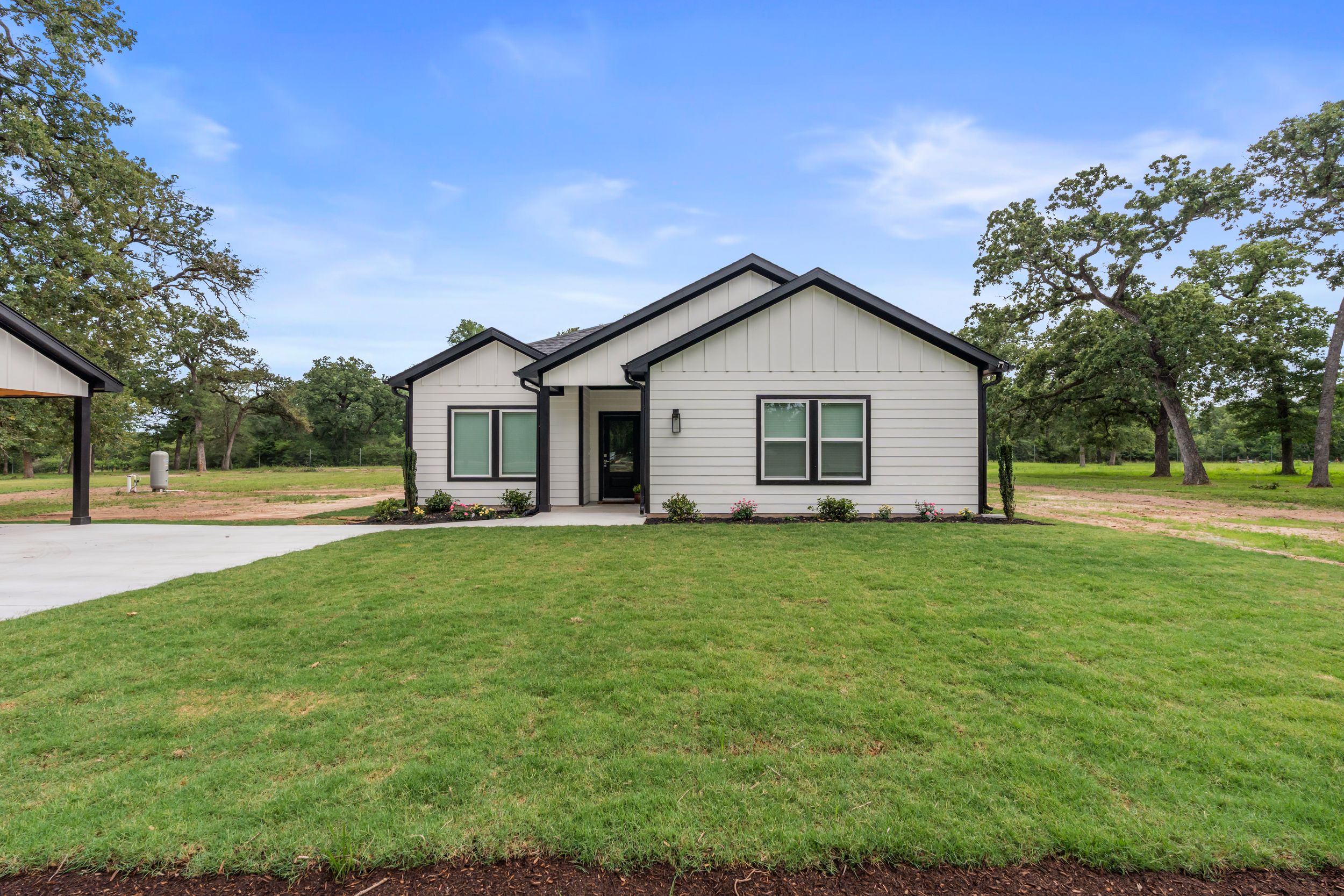 Modern white farmhouse with black trim, lush green lawn, and mature oak trees under a blue sky.