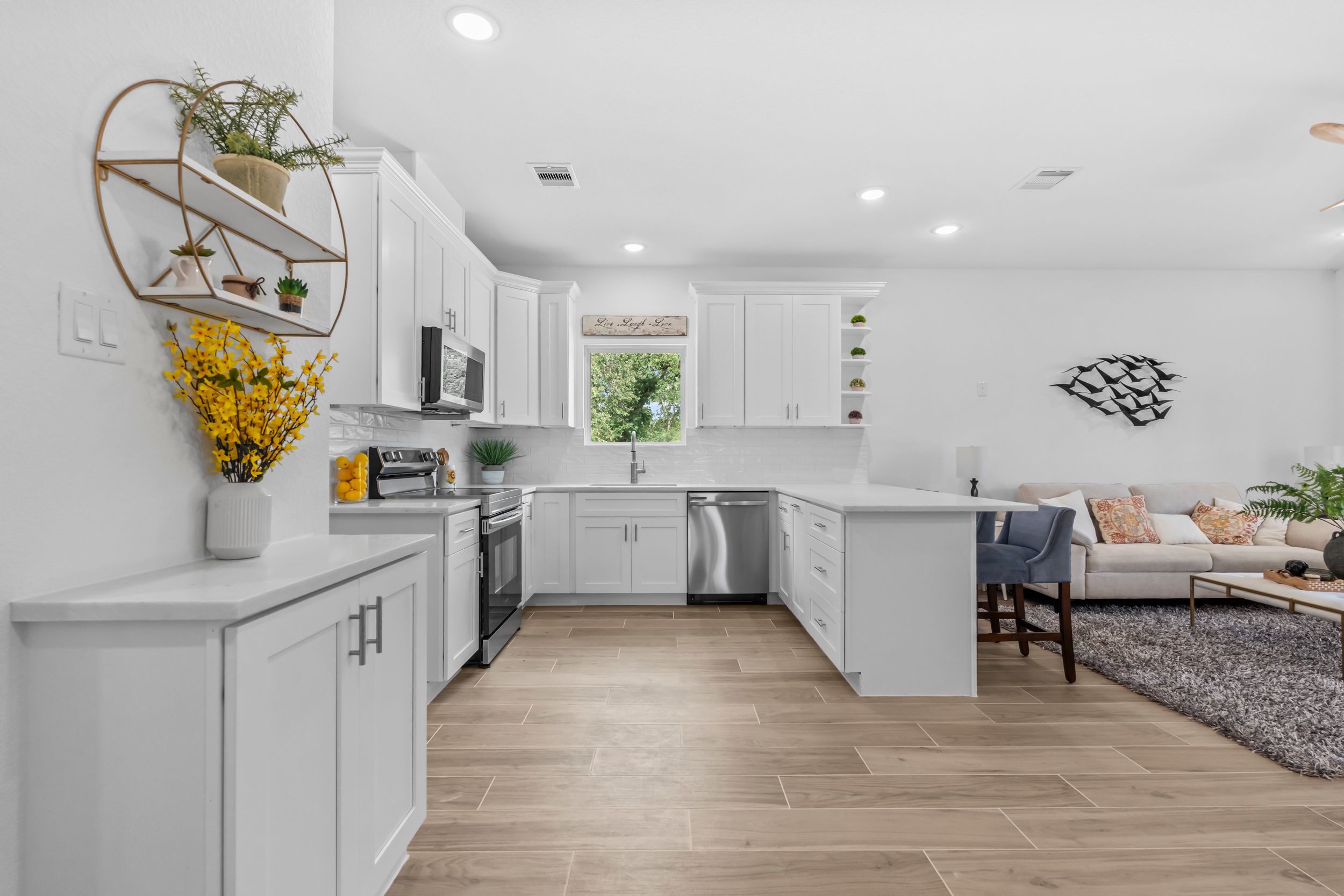 Modern white kitchen with stainless steel appliances, light wood floors, and open layout connecting to living area.