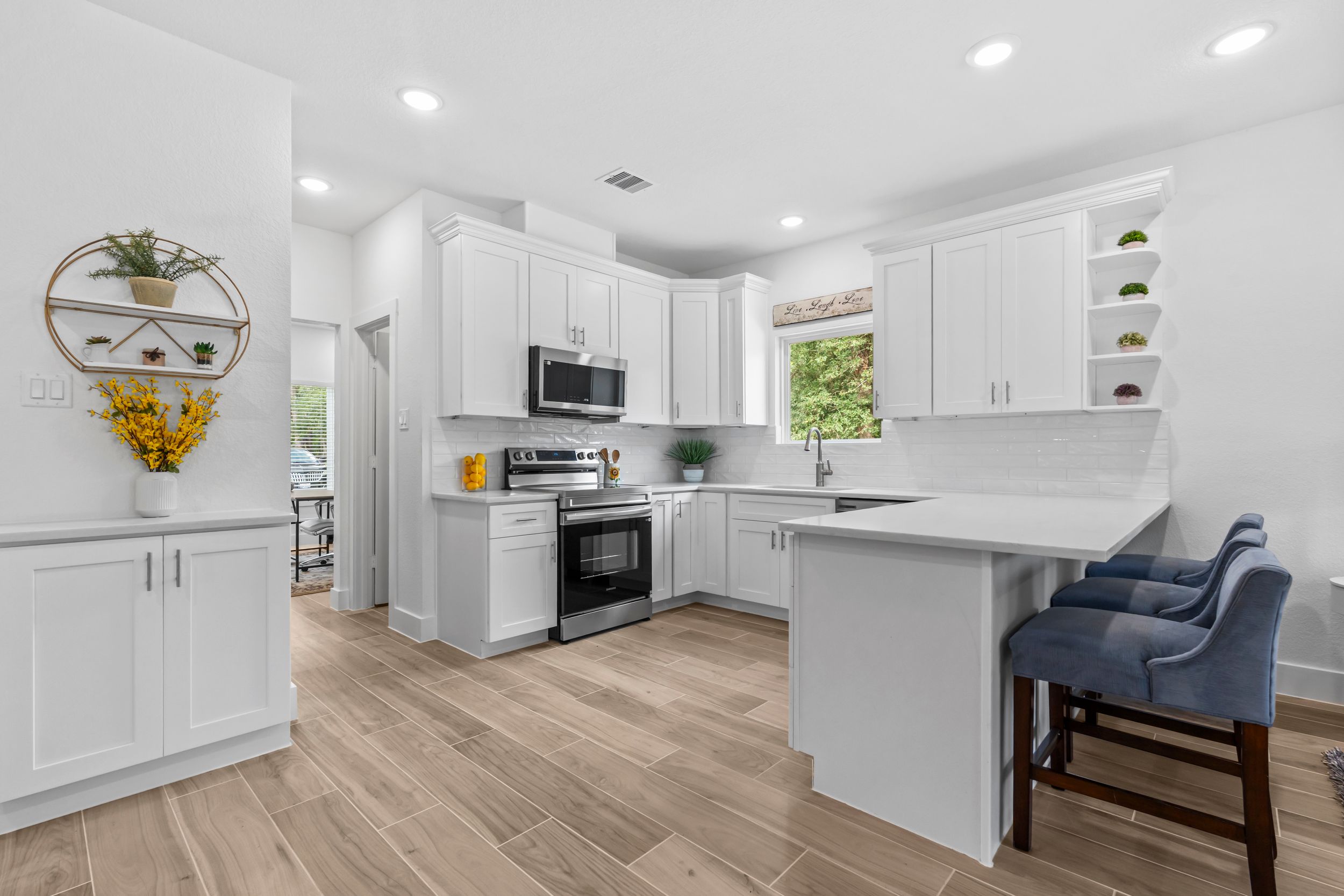 Bright white kitchen with stainless steel appliances, wood-look tile floors, open shelving, and a breakfast bar with blue chairs.