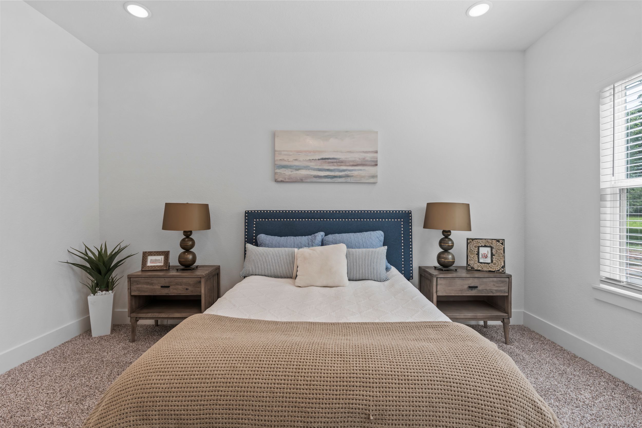 Cozy bedroom with blue upholstered headboard, neutral carpet, twin nightstands with lamps, and natural light from a window.