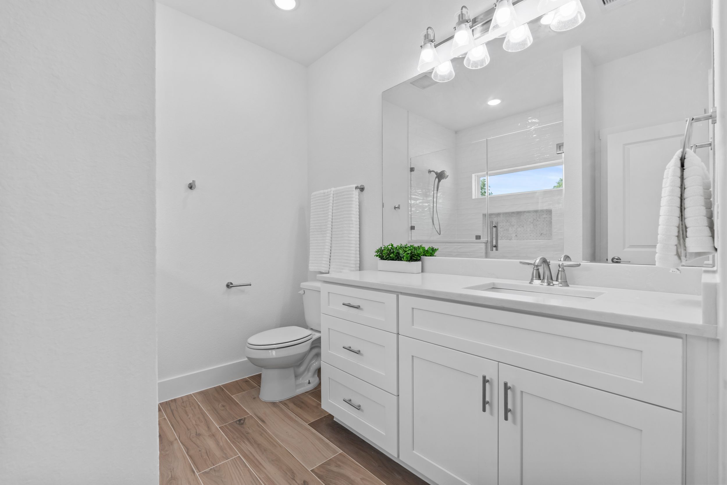 Modern white bathroom with double vanity, large mirror, wood-look tile floors, and bright lighting.