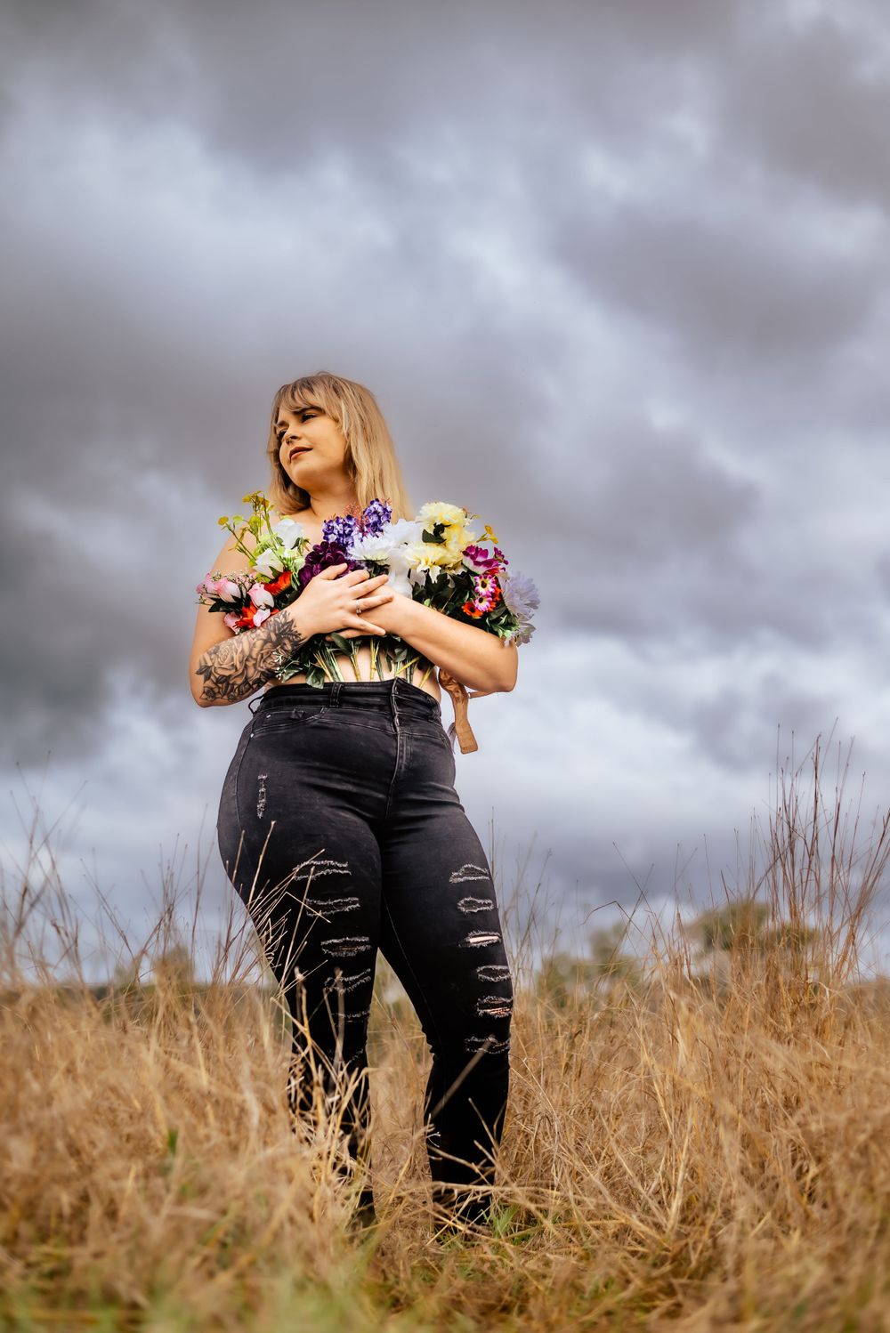 A dreamy portrait with vibrant flowers clutched to chest while standing in golden grass under stormy skies.