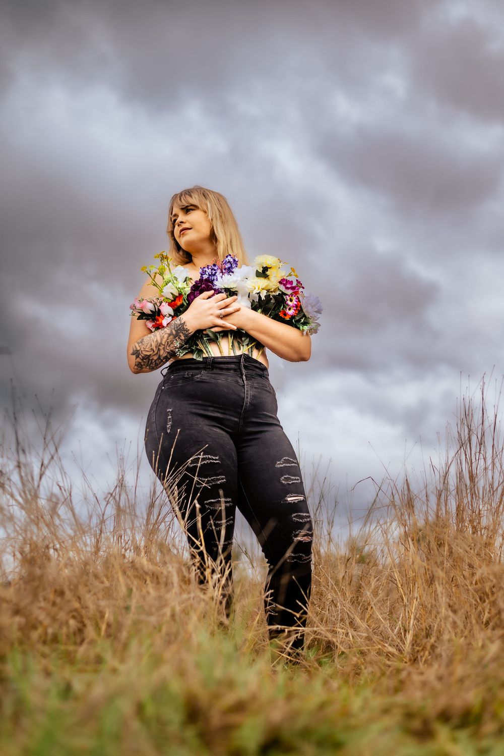 Standing figure holds vibrant wildflower bouquet in golden field under cloudy sky.