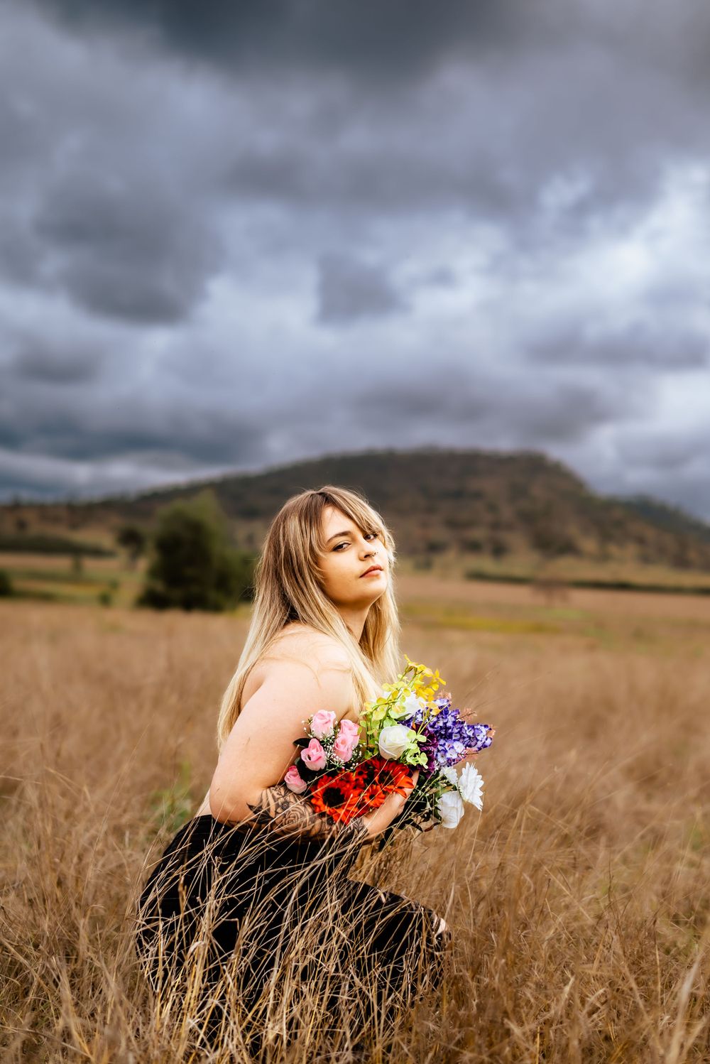 Artistic portrait with mixed flowers against dramatic cloudy sky and rural landscape backdrop.