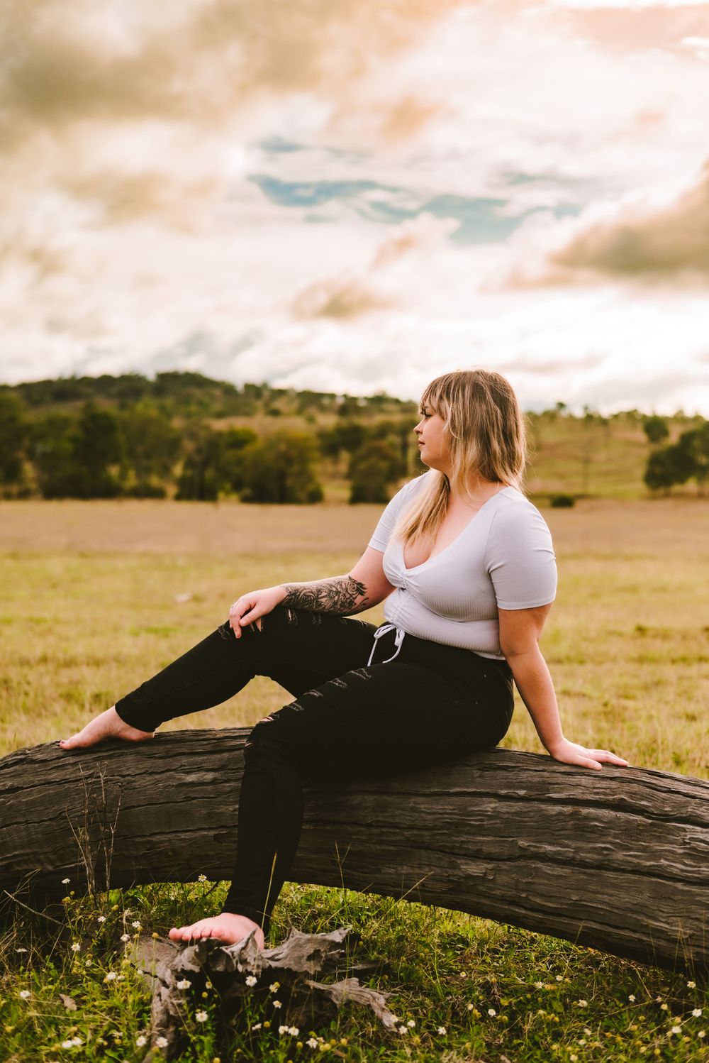 Relaxed outdoor portrait sitting on fallen log in sunlit field with mountains in background.