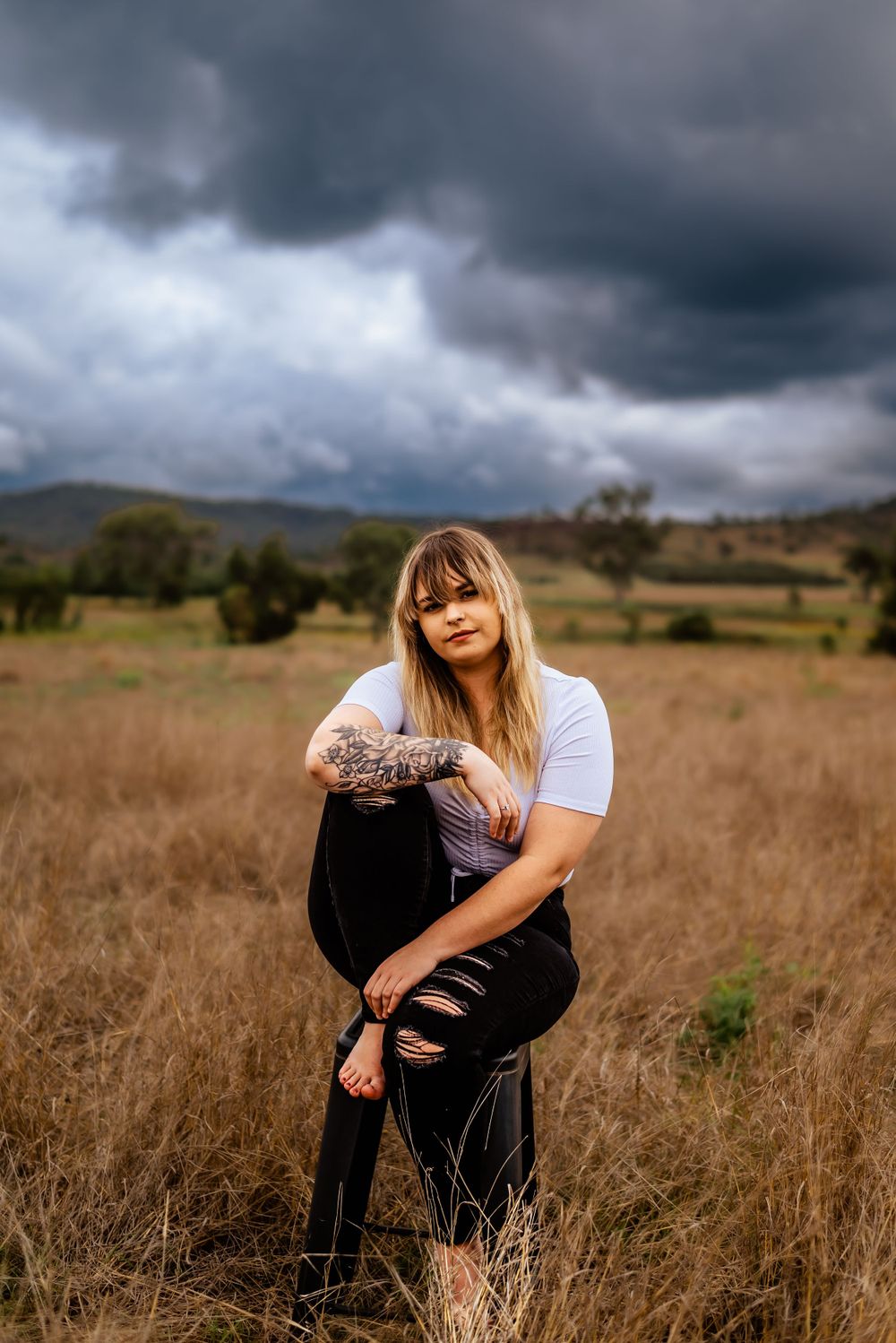 Tattooed figure poses thoughtfully in a rural field beneath dramatic storm clouds.
