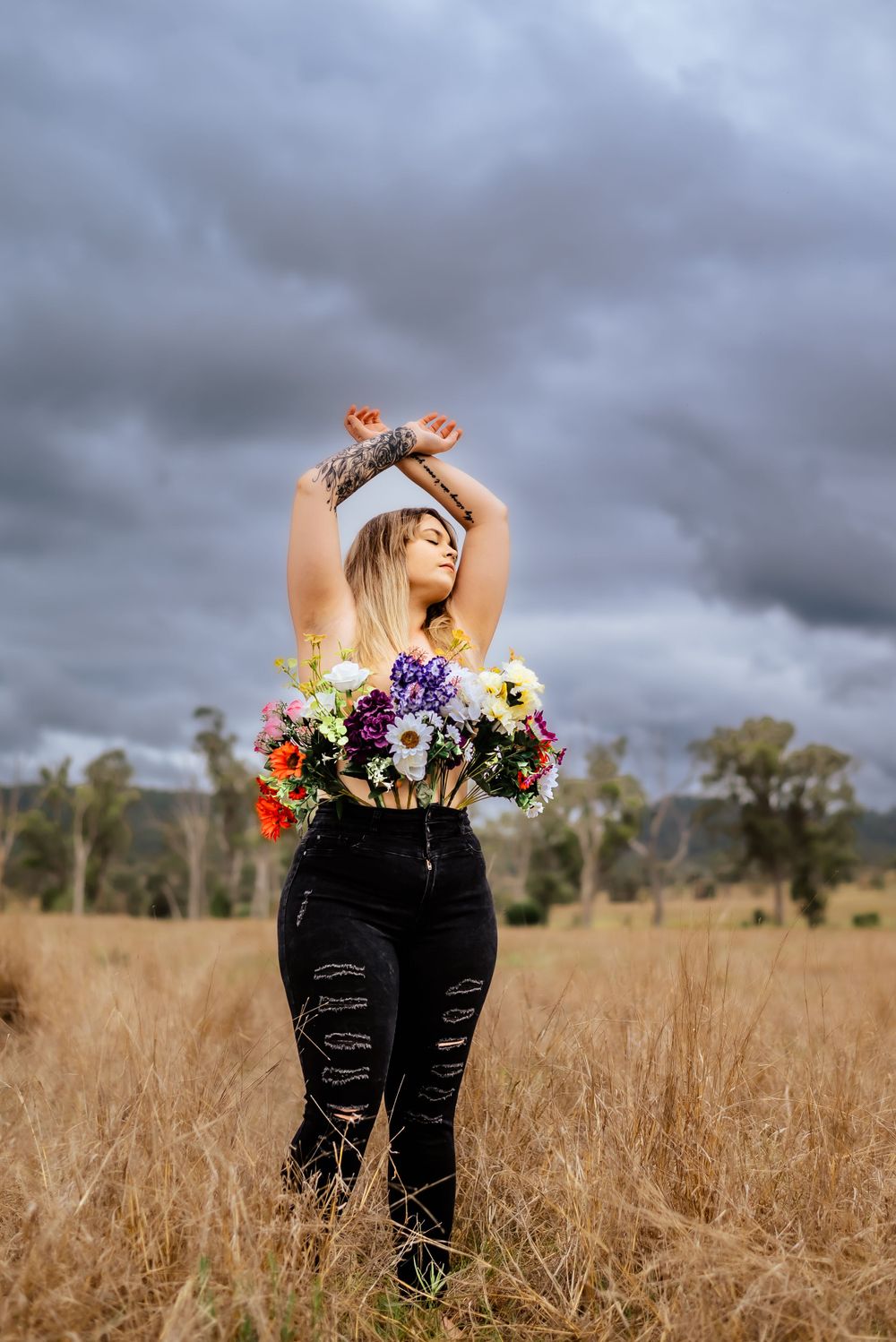 Artistic portrait with flowers and raised arms against stormy rural landscape.