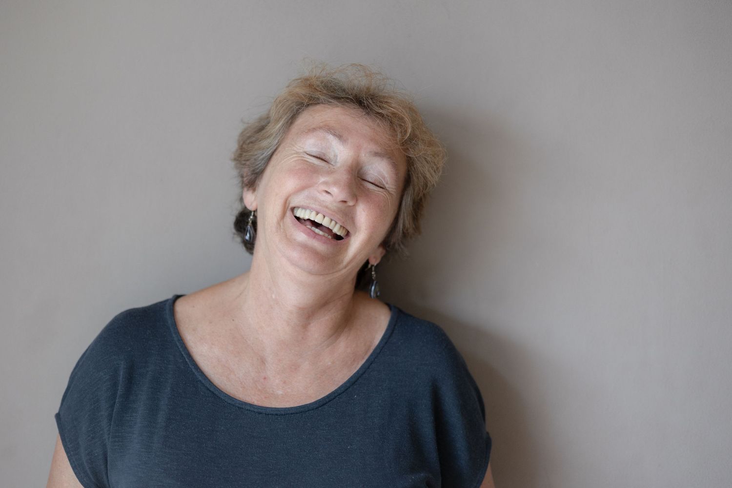 Candid portrait of someone laughing joyfully against a gray wall while wearing a dark top.