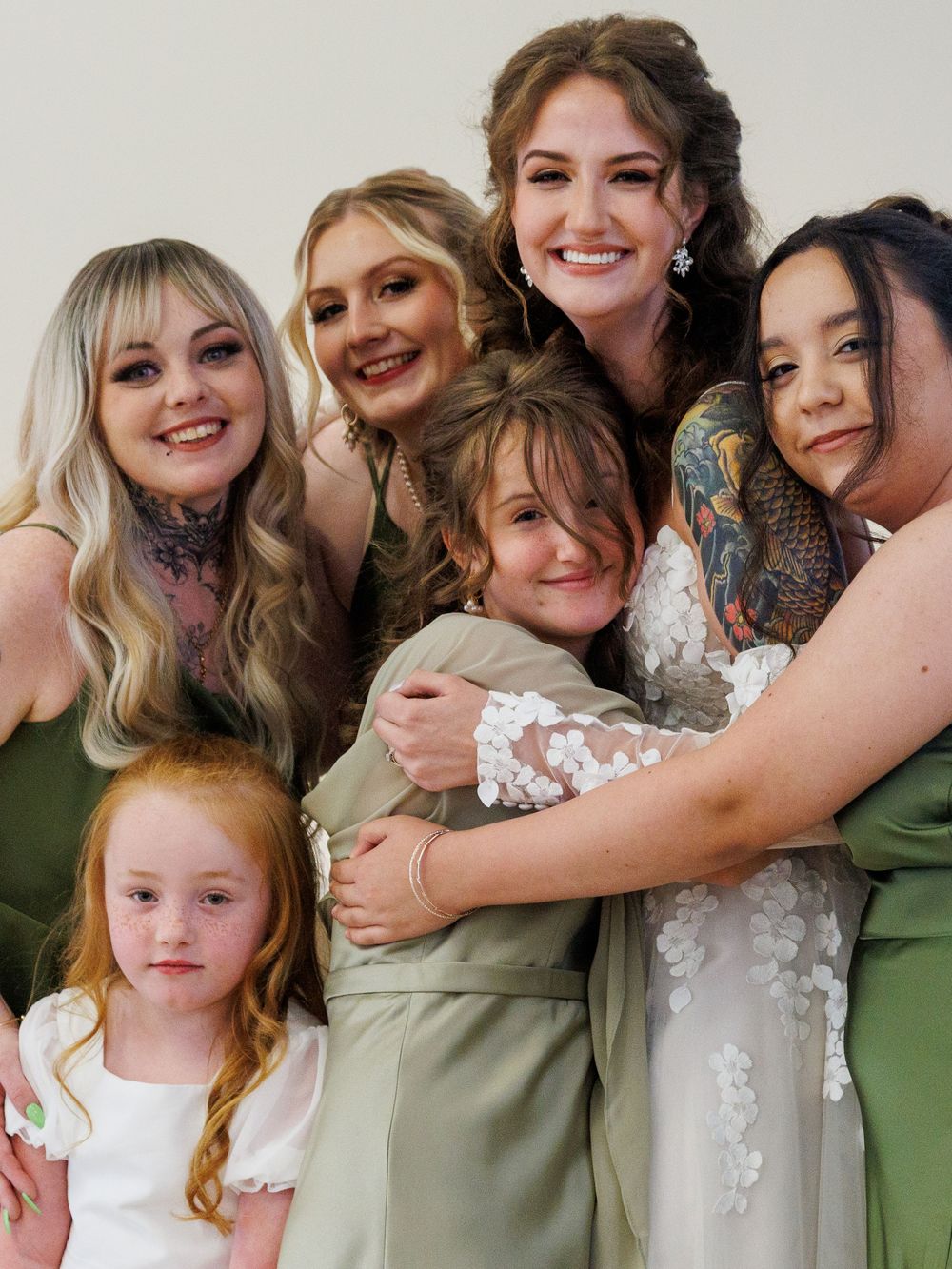 Bride in white lace gown hugging bridesmaids in sage green dresses with a flower girl at a wedding.