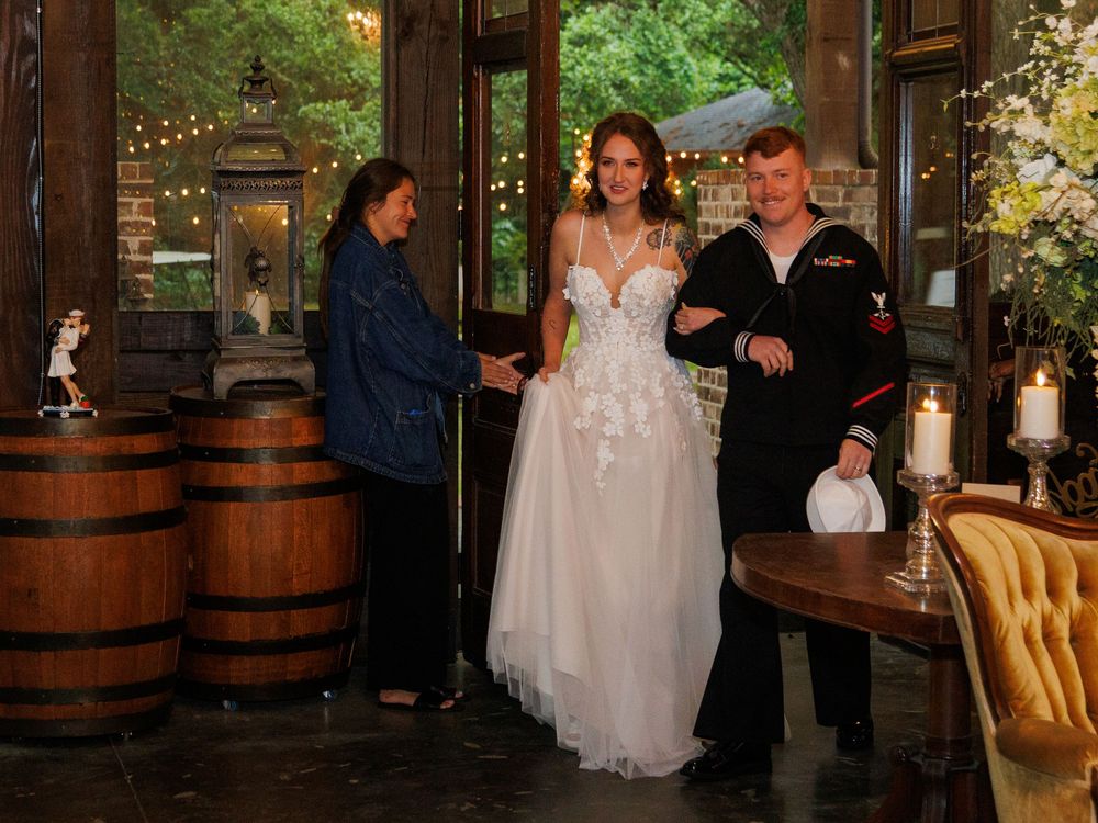 Navy sailor in uniform escorts bride in white gown through rustic venue with wooden barrels and string lights.