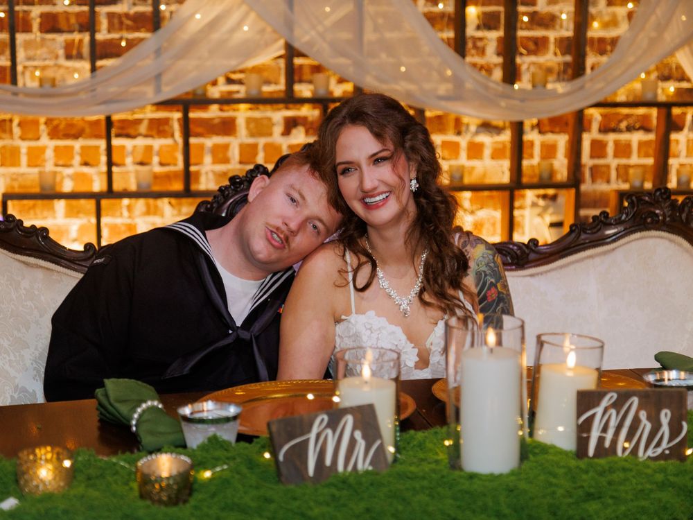 Newlywed couple smiles at sweetheart table with Mr. and Mrs. signs, candles, and greenery at their wedding reception.