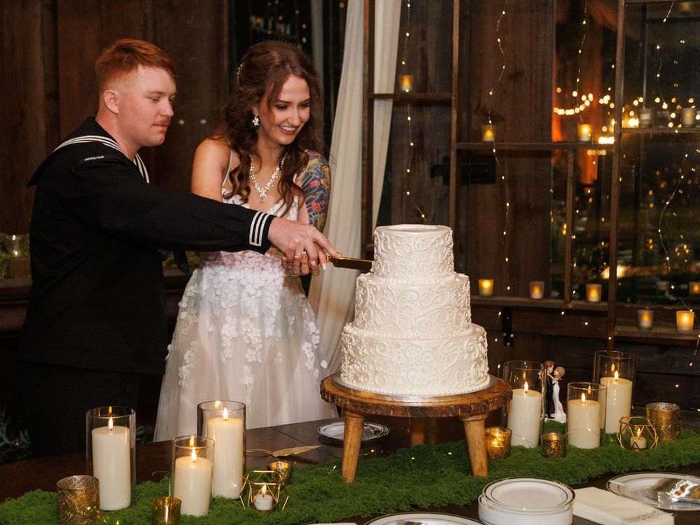 Navy sailor and bride cutting a white three-tier wedding cake surrounded by candles at a romantic reception.