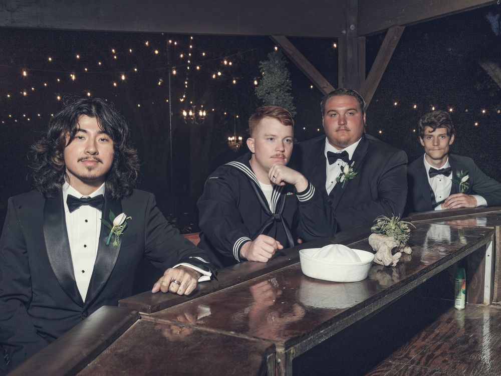 Four groomsmen in black tuxedos sitting at a rustic bar with string lights at a wedding reception.