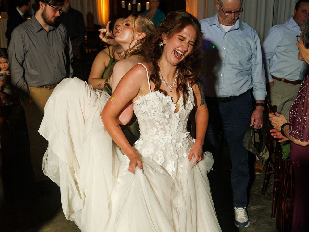 Two brides in white wedding gowns laughing and dancing joyfully at a wedding reception celebration.