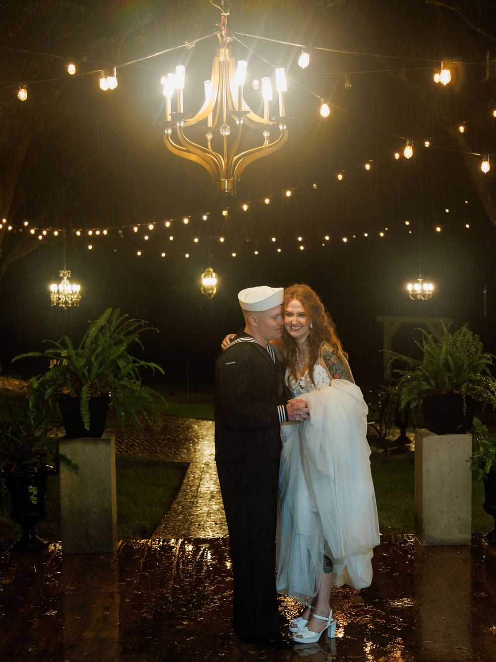 Navy sailor and bride share romantic dance under chandelier and string lights at elegant outdoor wedding reception.