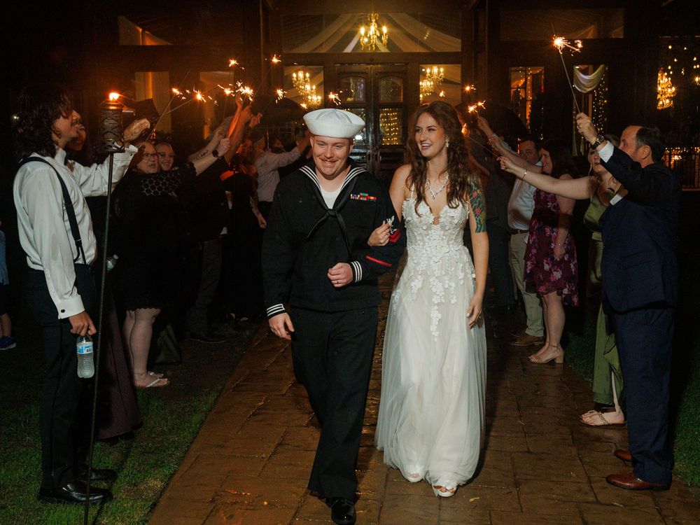 Navy sailor and bride exit wedding reception through sparkler tunnel with cheering guests at night.