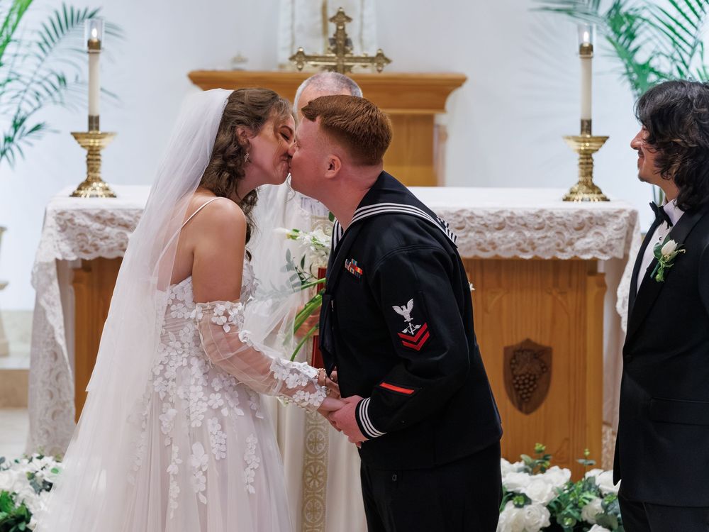 Navy sailor groom kisses bride in white lace wedding dress at church altar ceremony.
