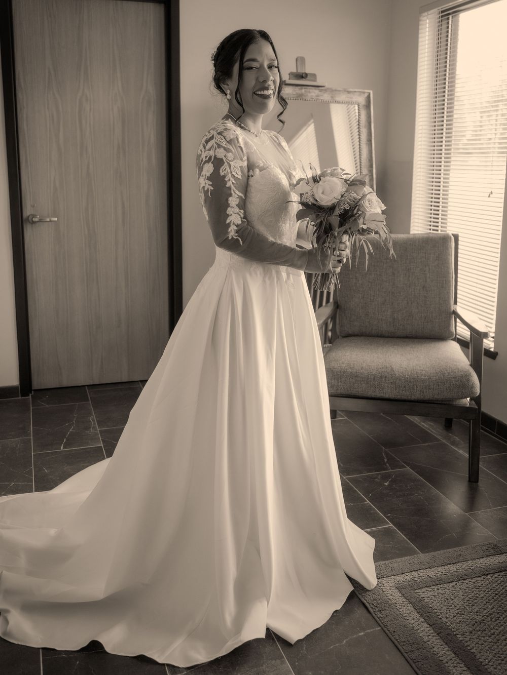 A bride in an elegant long-sleeved white wedding gown holds a bouquet while standing in a room, captured in black and white.