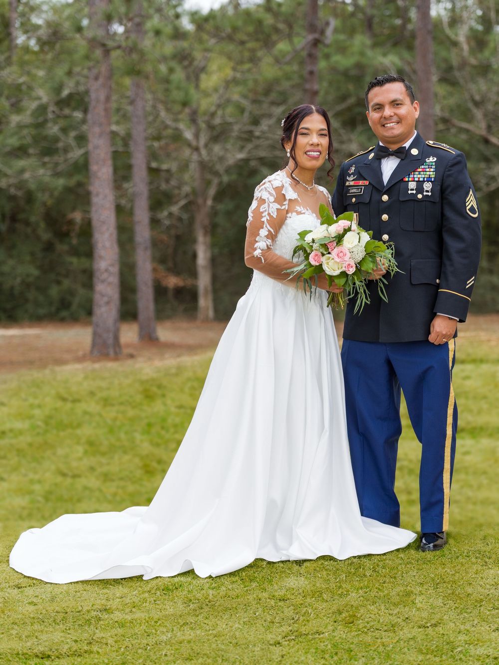 A couple poses together on a grassy lawn, with the groom in military dress uniform and bride in a flowing white gown.