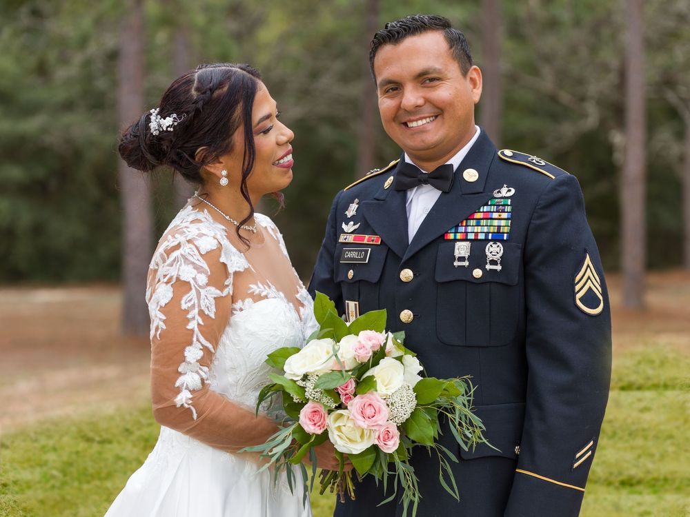 A bride in a white lace gown holds a pink and white bouquet while standing with a uniformed service member outdoors.