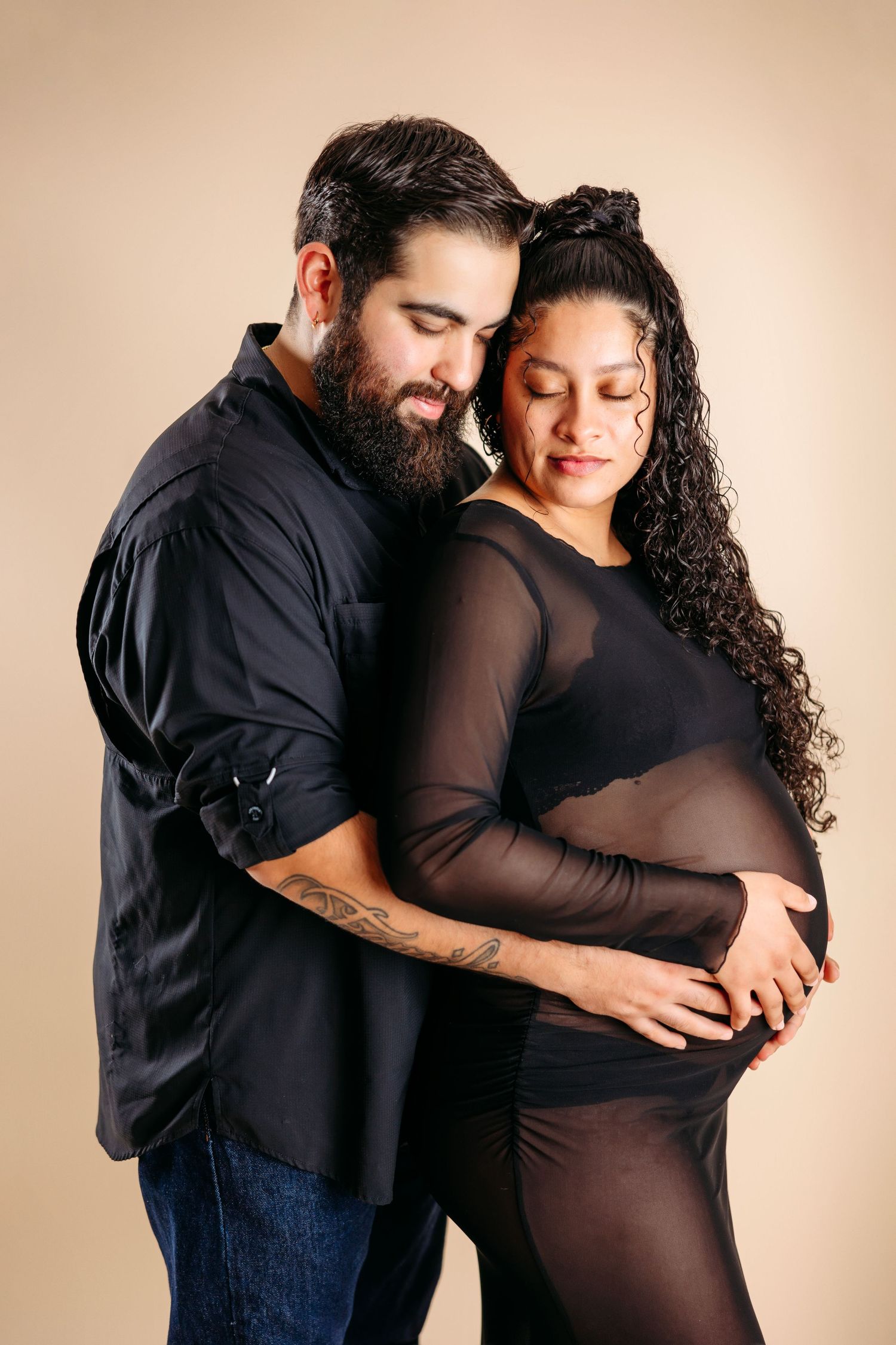 A couple in black clothing share an intimate moment during a maternity photoshoot against a beige backdrop. Maternity Photographer