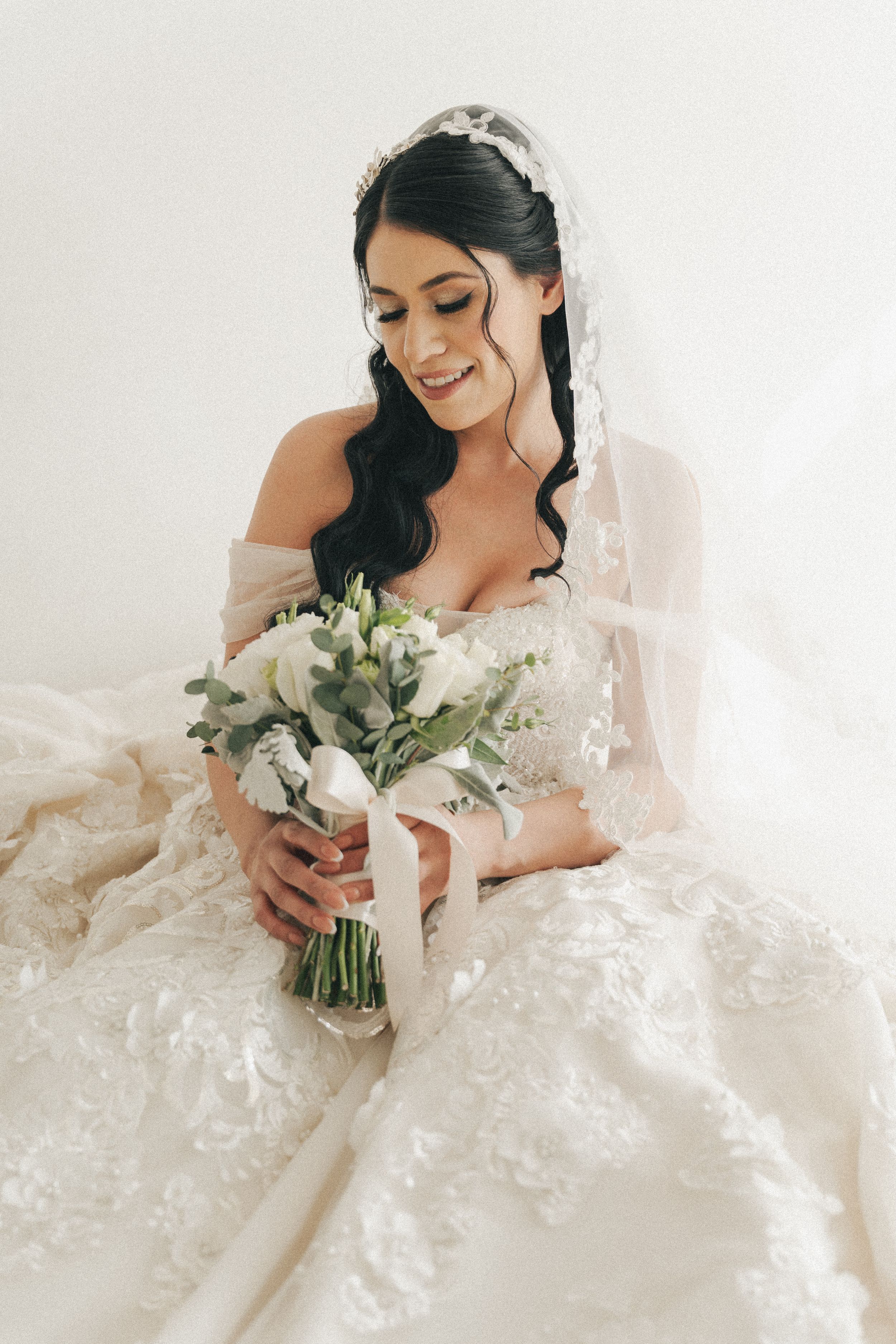 Elegant bride in lace wedding gown holding white floral bouquet against light background.
