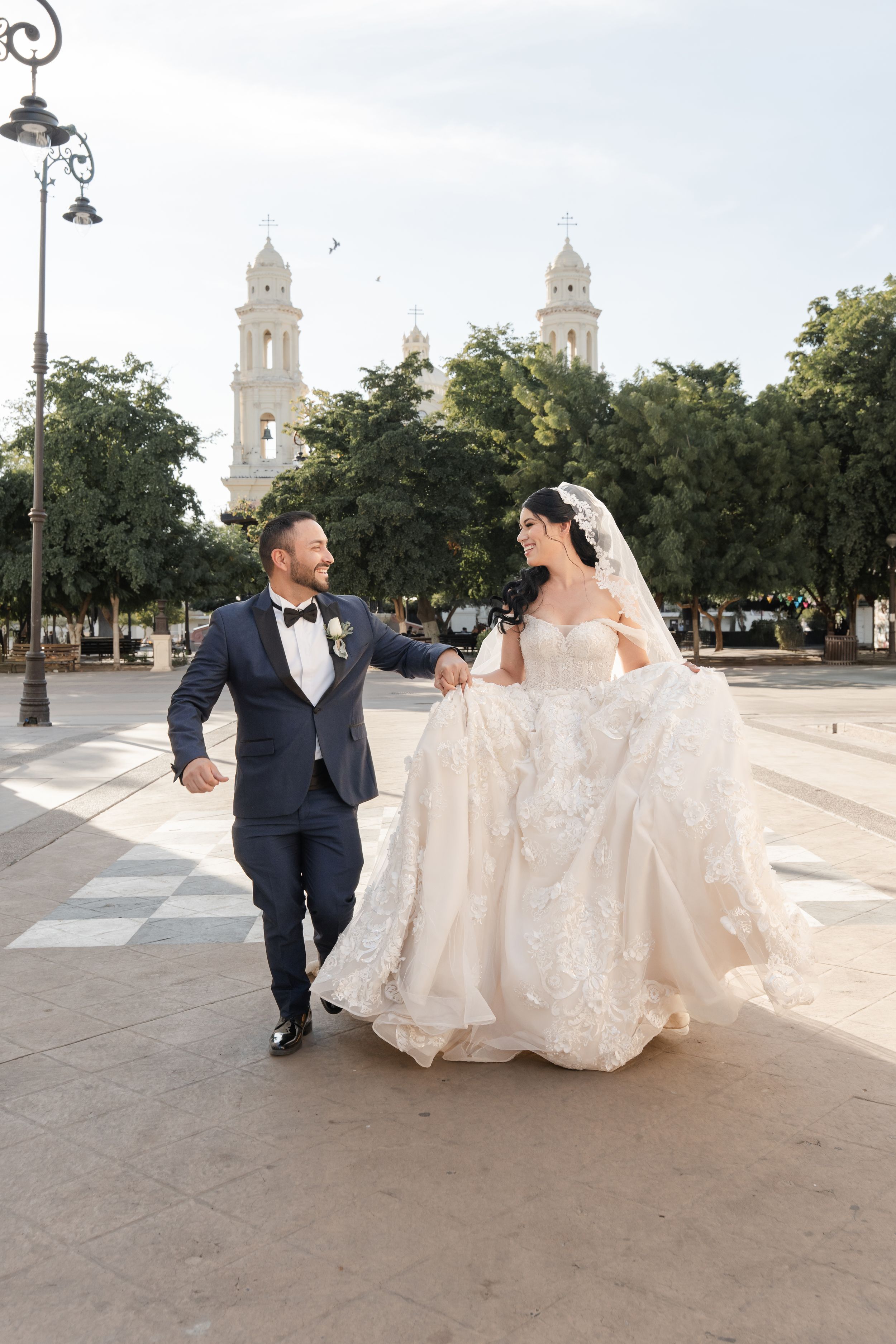 Newlyweds in formal attire walking and posing together on a sunny plaza with architectural backdrop.