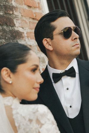 Stylish wedding couple in formal attire and sunglasses against brick wall.