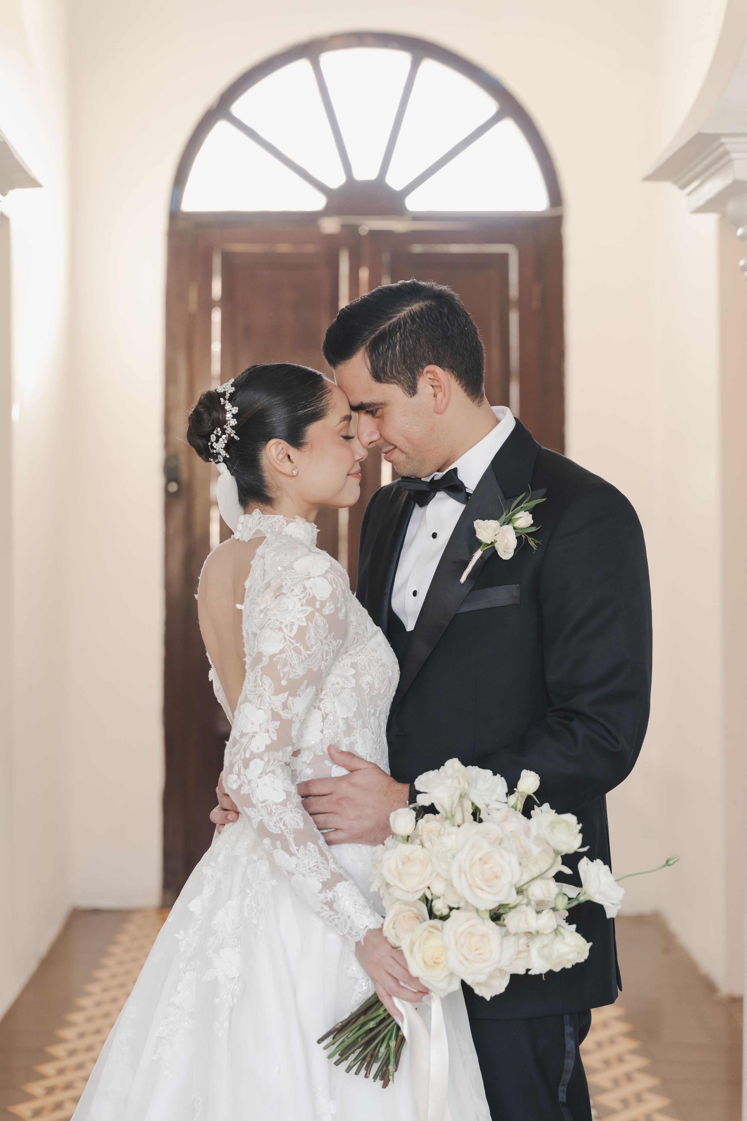 Elegant wedding couple in traditional attire sharing romantic moment in bright hallway.