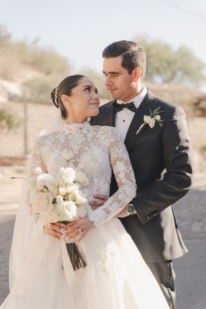 Wedding couple in formal attire embracing outdoors in desert setting with white bouquet.
