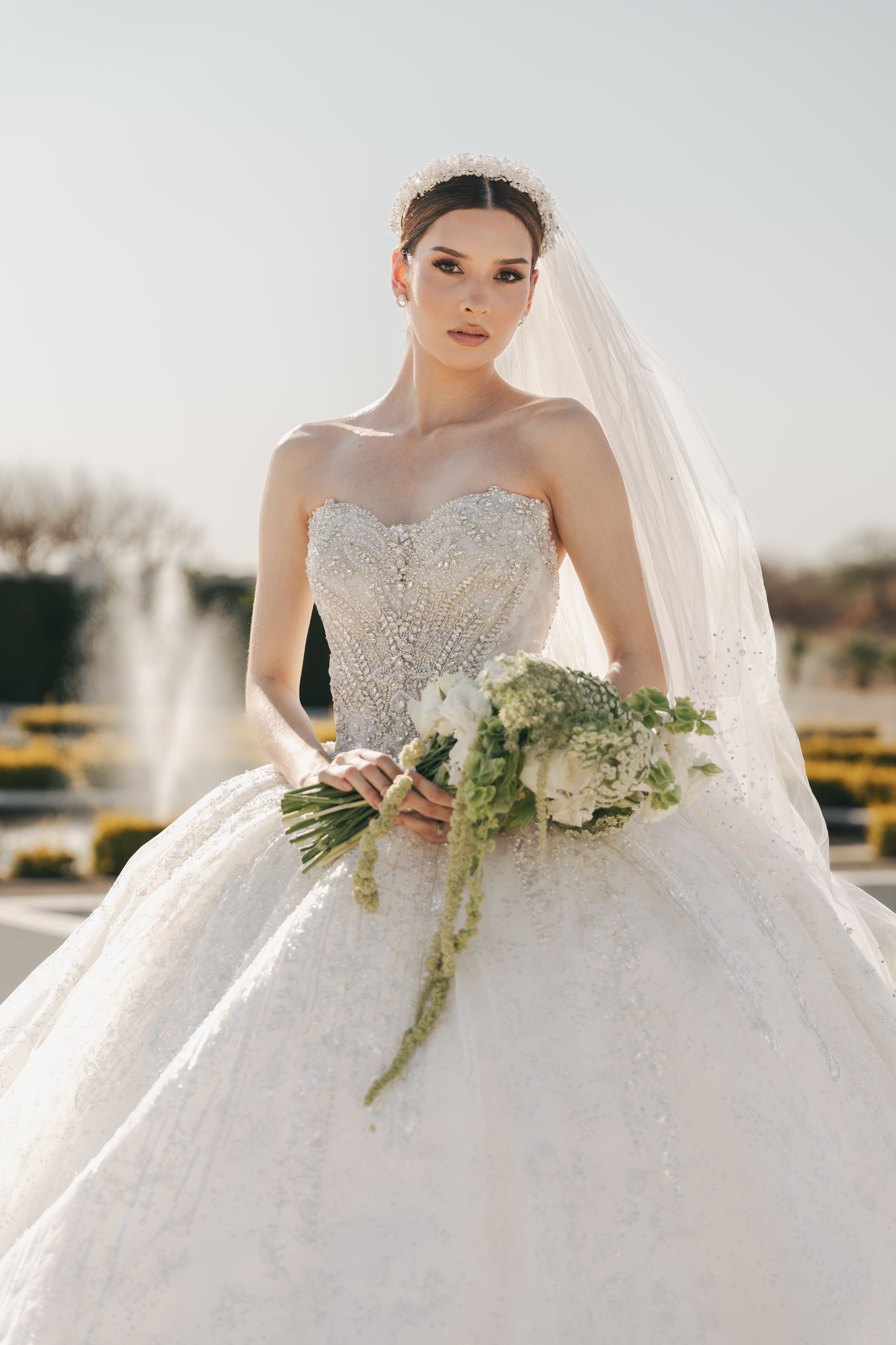 Bride in beaded ballgown holding cascading bouquet against light background.