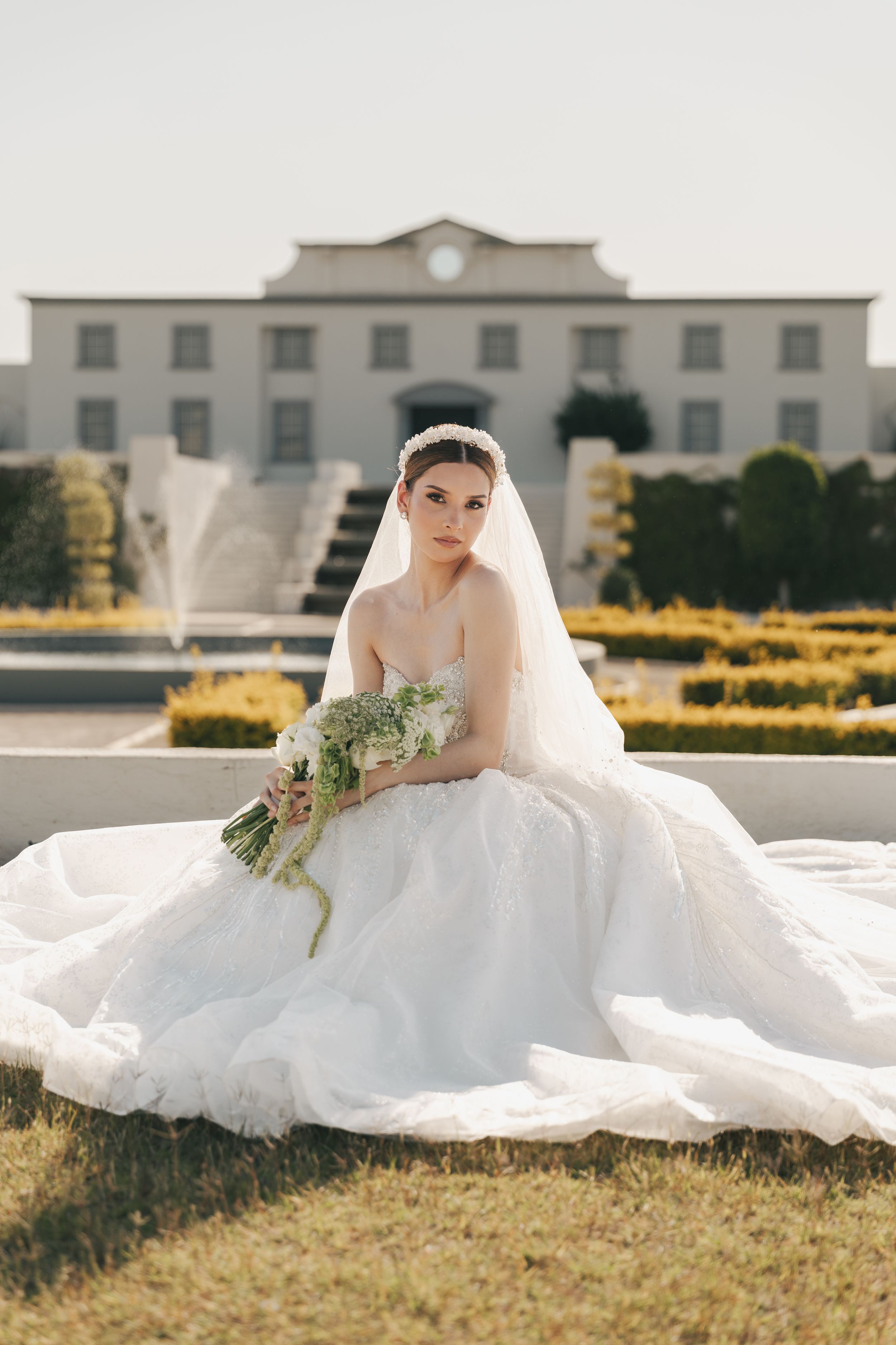 Bride in full ballgown wedding dress posed on manicured lawn with historic building backdrop.