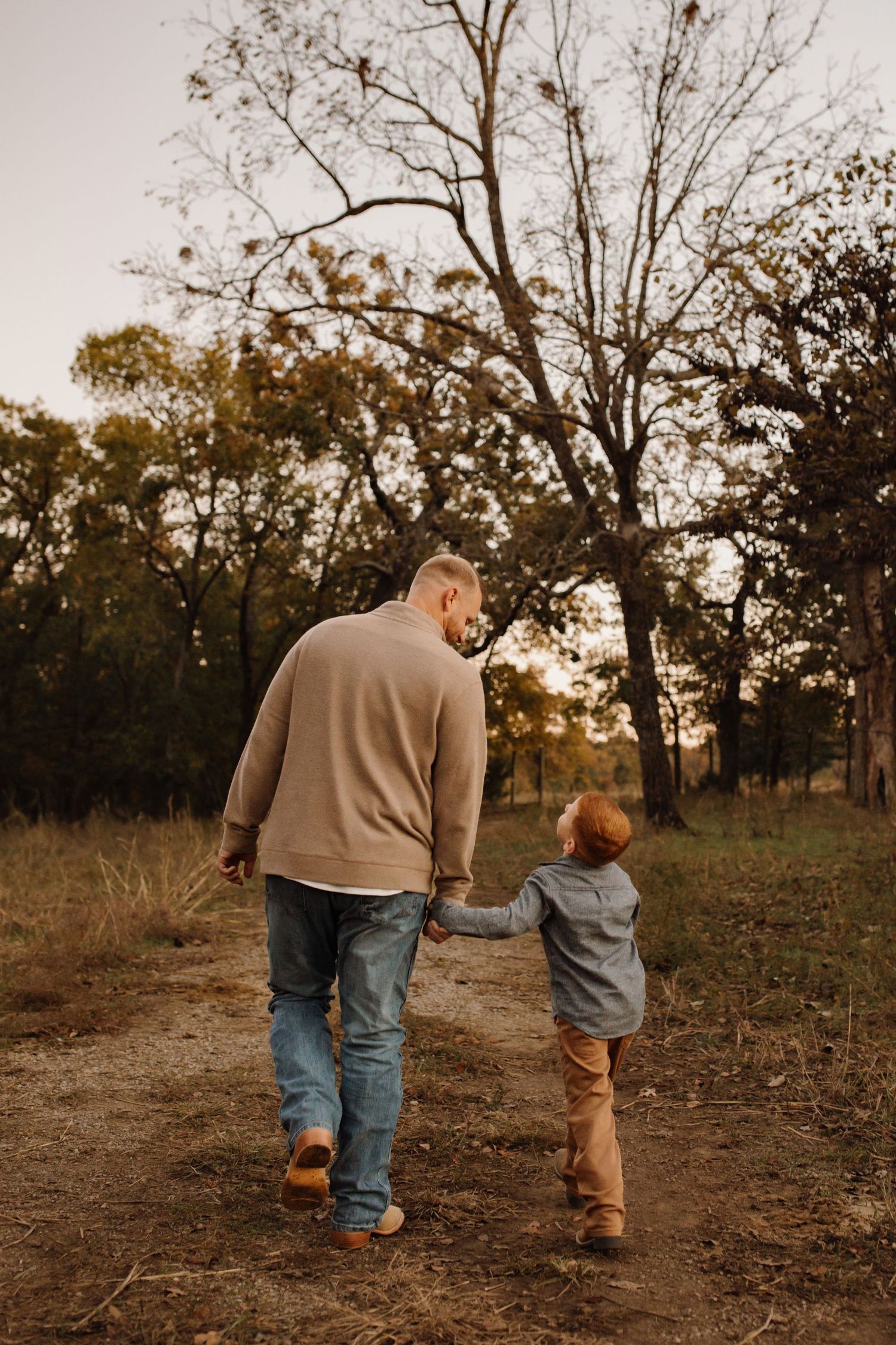Two people walk hand in hand down a dirt path at sunset through a wooded area on an autumn evening.