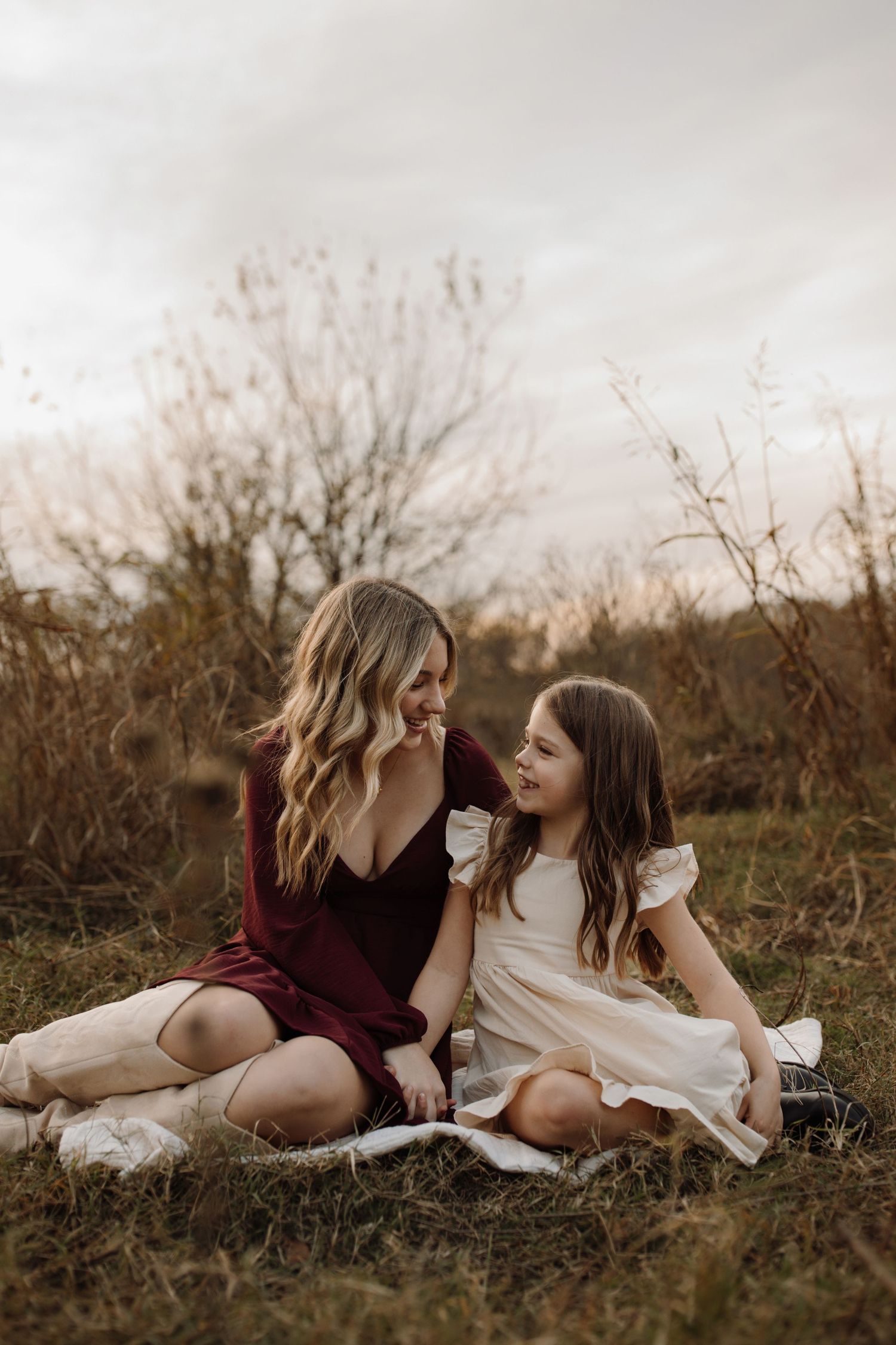 Two people in contrasting burgundy and white dresses lay together in a grassy field at sunset.