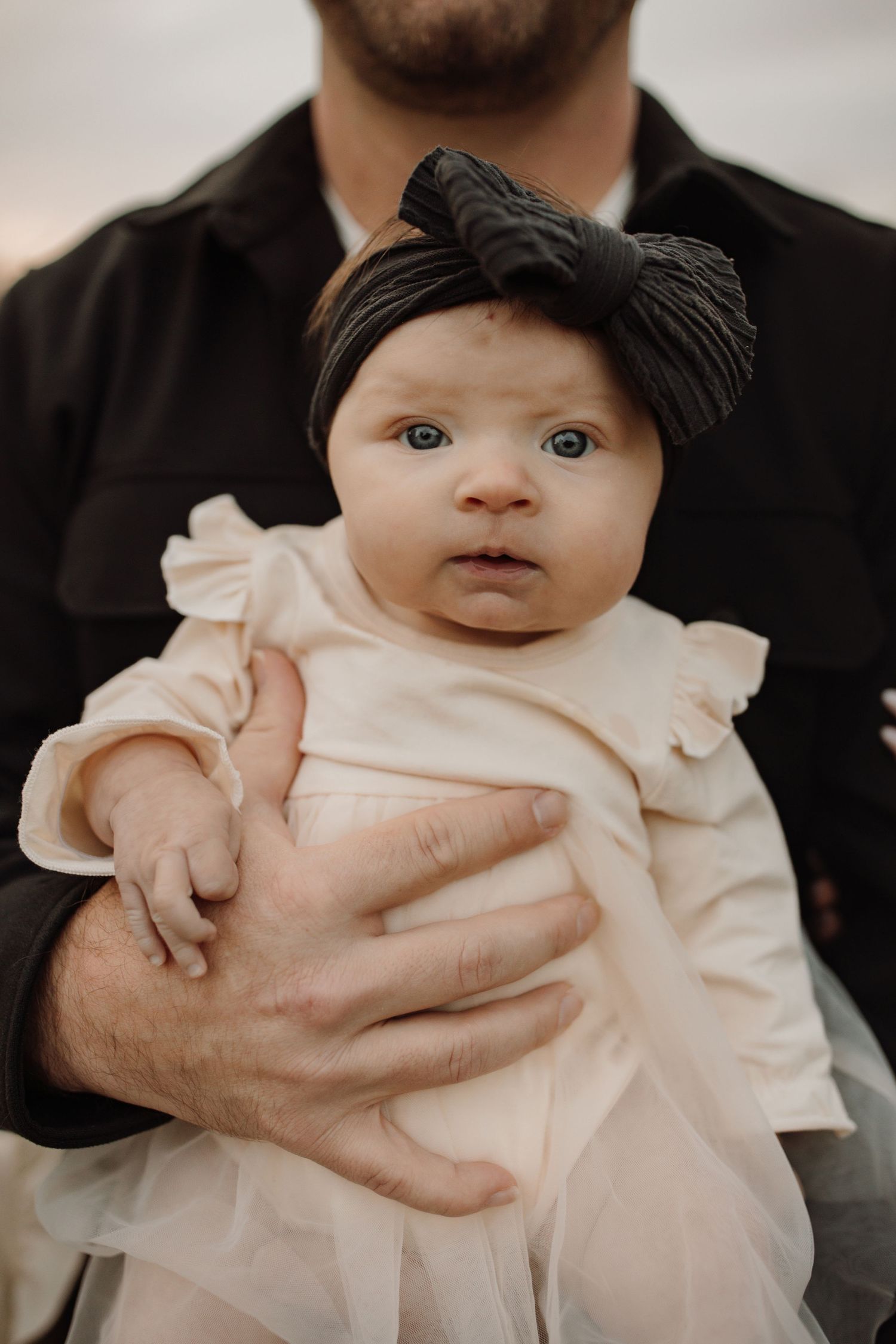 Baby wearing black bow headband and cream ruffled dress is held by someone in black clothing.