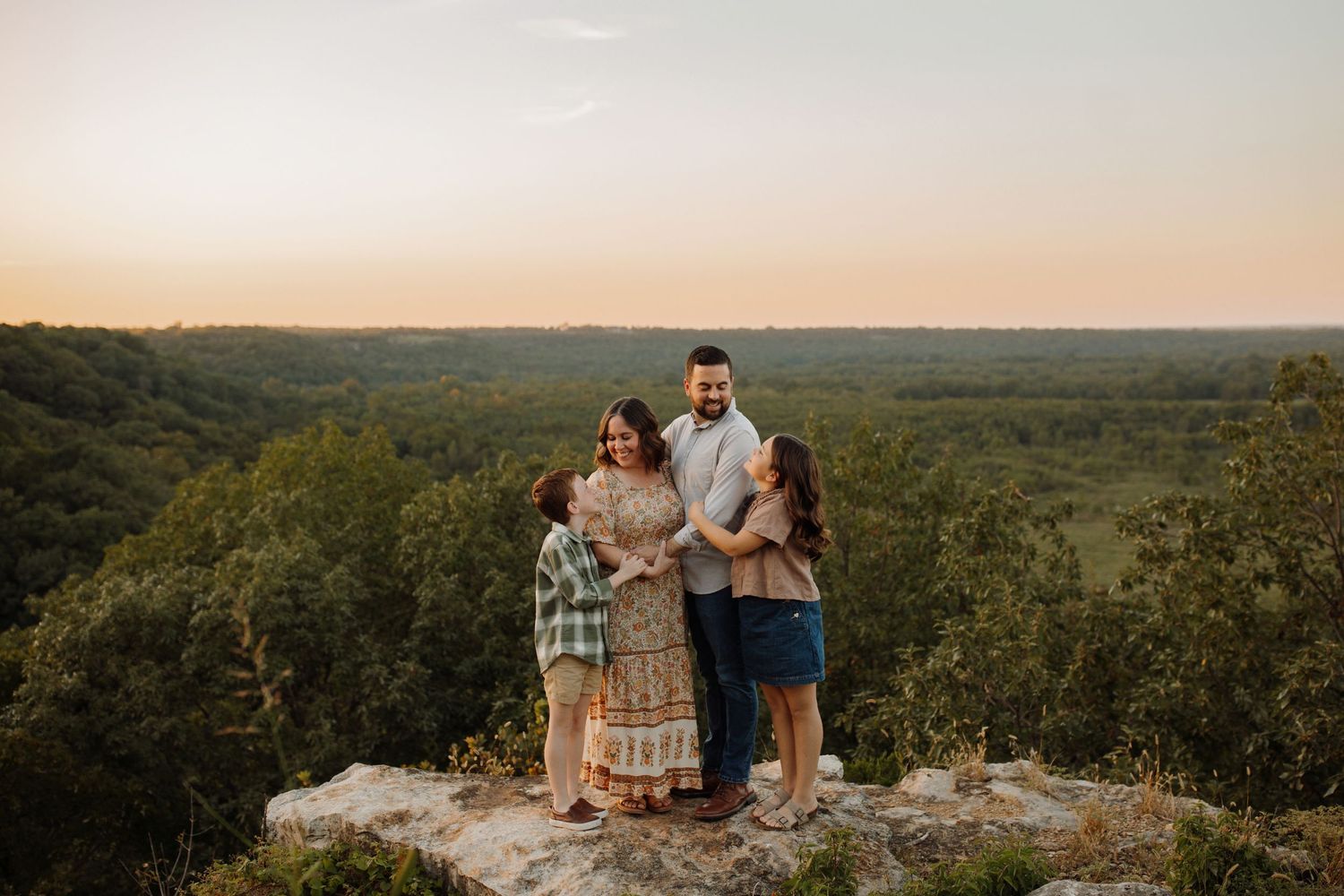 A family stands together on a rocky overlook during sunset with a vast forested landscape stretching into the horizon.