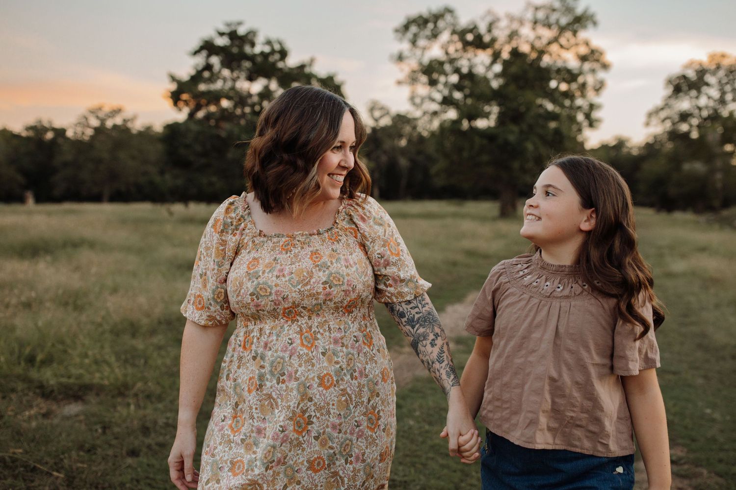 Two people in floral dresses stand together in a sunlit grassy field during golden hour.