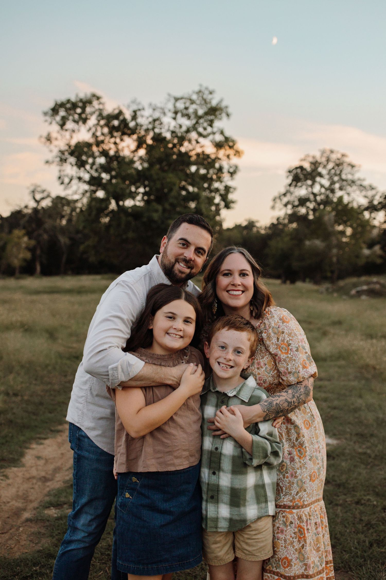 A family poses together outdoors at sunset in a grassy field with trees silhouetted in the background.