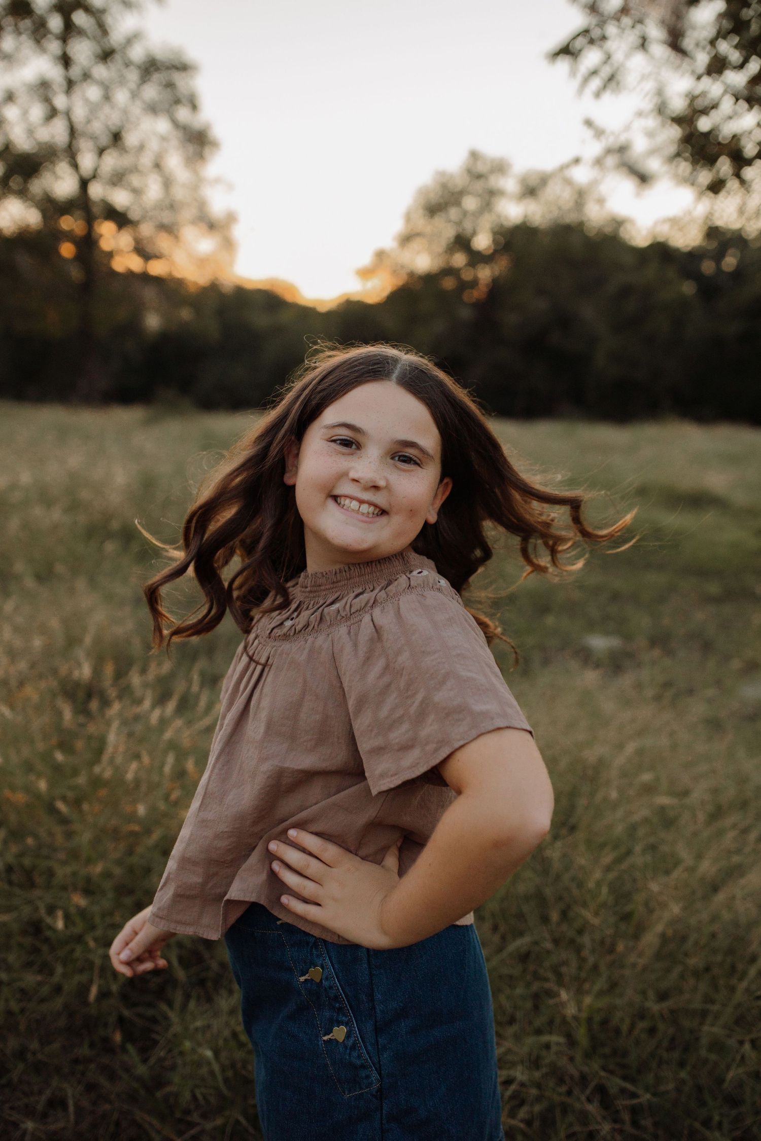 A person in a brown top and jeans twirls playfully in a sunlit field at golden hour.